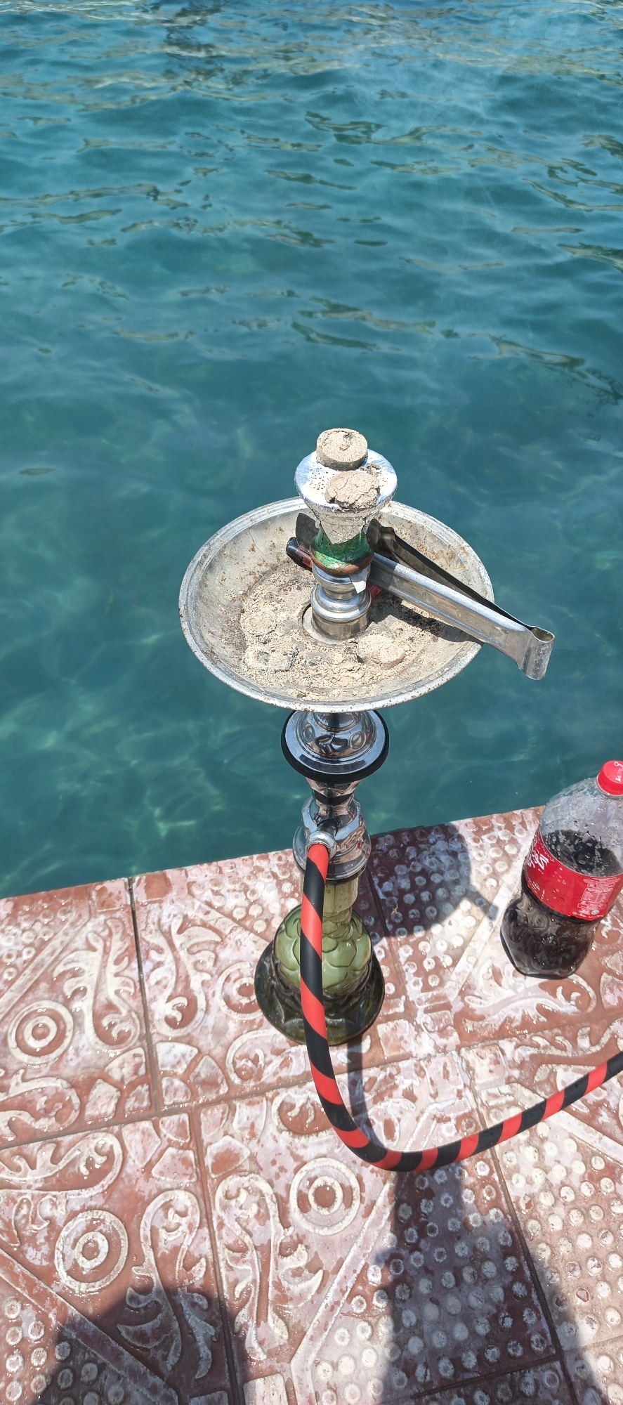 A high-angle outdoor shot captures a hookah and a Coca-Cola bottle positioned on patterned tiles next to a clear, deep blue body of water, possibly a sea or large lake, under bright daylight.
In the foreground, a silver-colored hookah with a green glass base and a red and black striped hose stands on reddish-brown tiles. The tiles feature intricate raised patterns in white. The top tray of the hookah is visibly used, containing ash and remnants of coals, with metal tongs resting on it. On the right, a clear plastic Coca-Cola bottle with a red label shows condensation, suggesting it is cold. The brand name "Coca-Cola" is partially visible on the label.
The background is dominated by the serene, transparent blue-green water, showing ripples and reflections of sunlight, indicating calm conditions. The water's clarity reveals hints of the bottom or submerged features. Strong shadows cast by the hookah and bottle suggest a sunny day, likely midday or early afternoon. The scene conveys a relaxed outdoor setting by the water. The location context provided is Samundri, Pakistan.