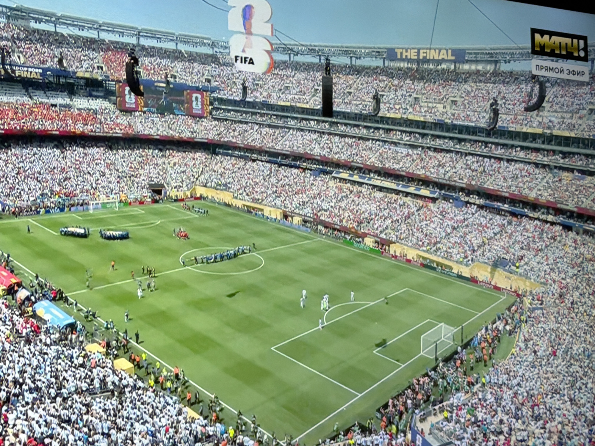 The image depicts a wide aerial view of a crowded football stadium, likely during a major sporting event given the signage "THE FINAL" and "WORLD CUP 2022". The stadium is filled with spectators, many wearing white and red attire. On the field, players are visible, seemingly during a pre-game ceremony or warm-up, as two teams are lined up on opposite sides of the pitch. A large screen in the background displays "22 23 FIFA". Another screen to the right shows "MATH!" and "ПРЯМОЙ ЭФИР" (Live Broadcast in Russian). The stadium architecture suggests a modern design with multiple tiers of seating. The overall atmosphere appears to be festive and energetic, characteristic of a championship match. There is no visible indication of the city of Orenburg, Russia in this image.