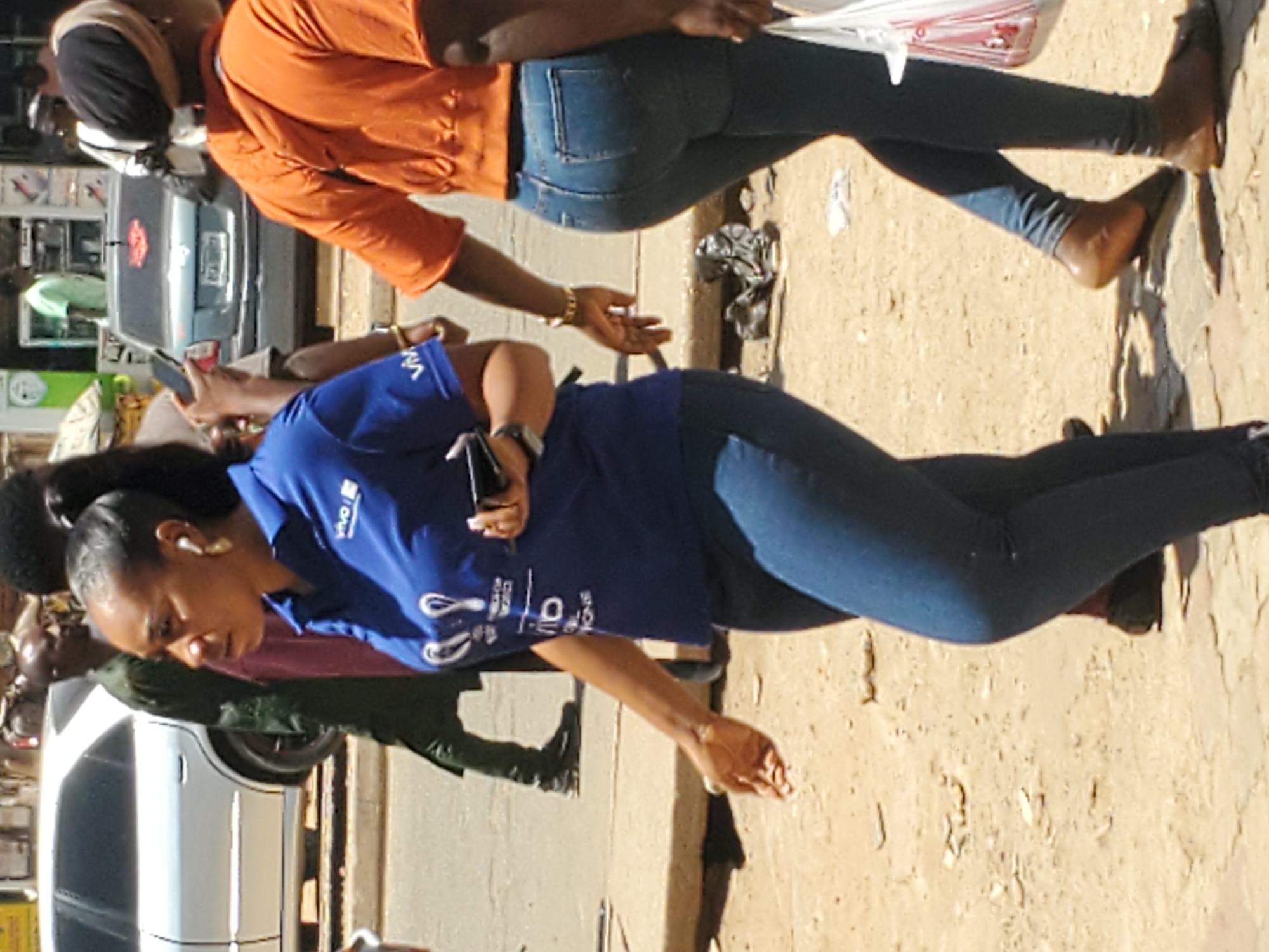The image depicts two women walking on a street in Jos, Nigeria. The woman in the foreground wears a blue shirt, blue jeans, and has a ponytail. She is looking down and appears to be holding a phone. She also has an earpiece in her ear. The woman in the background wears an orange top and blue jeans. She is carrying a plastic bag and wearing sandals. Both women are facing right, away from the camera. The ground is a light brown/tan color, and there are vehicles parked on the street. Street signage can be seen in the background.