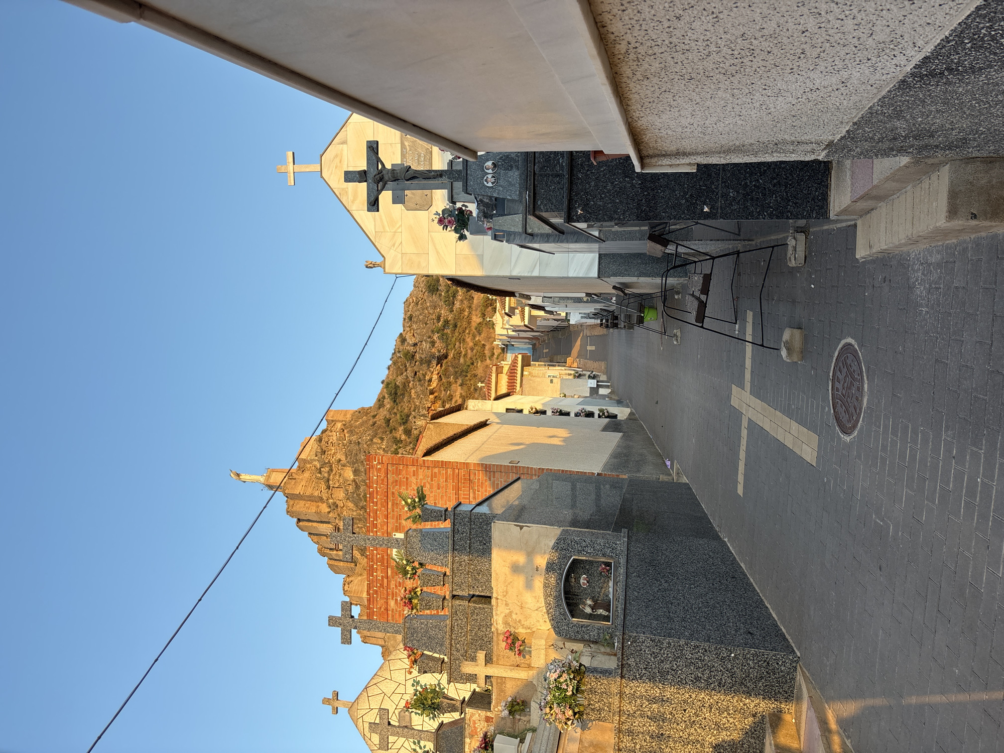 This is a photograph taken in a cemetery located in the city of Murcia, Spain. The image captures a narrow pathway lined with various tombs and mausoleums, all of which are adorned with Christian crosses. The pathway is paved with dark gray bricks, and a yellow cross made of bricks is embedded in the pavement. In the background, a statue of Jesus Christ is visible atop a rocky hill, with a clear blue sky overhead. The lighting suggests that it is either early morning or late afternoon, casting long shadows across the scene.
