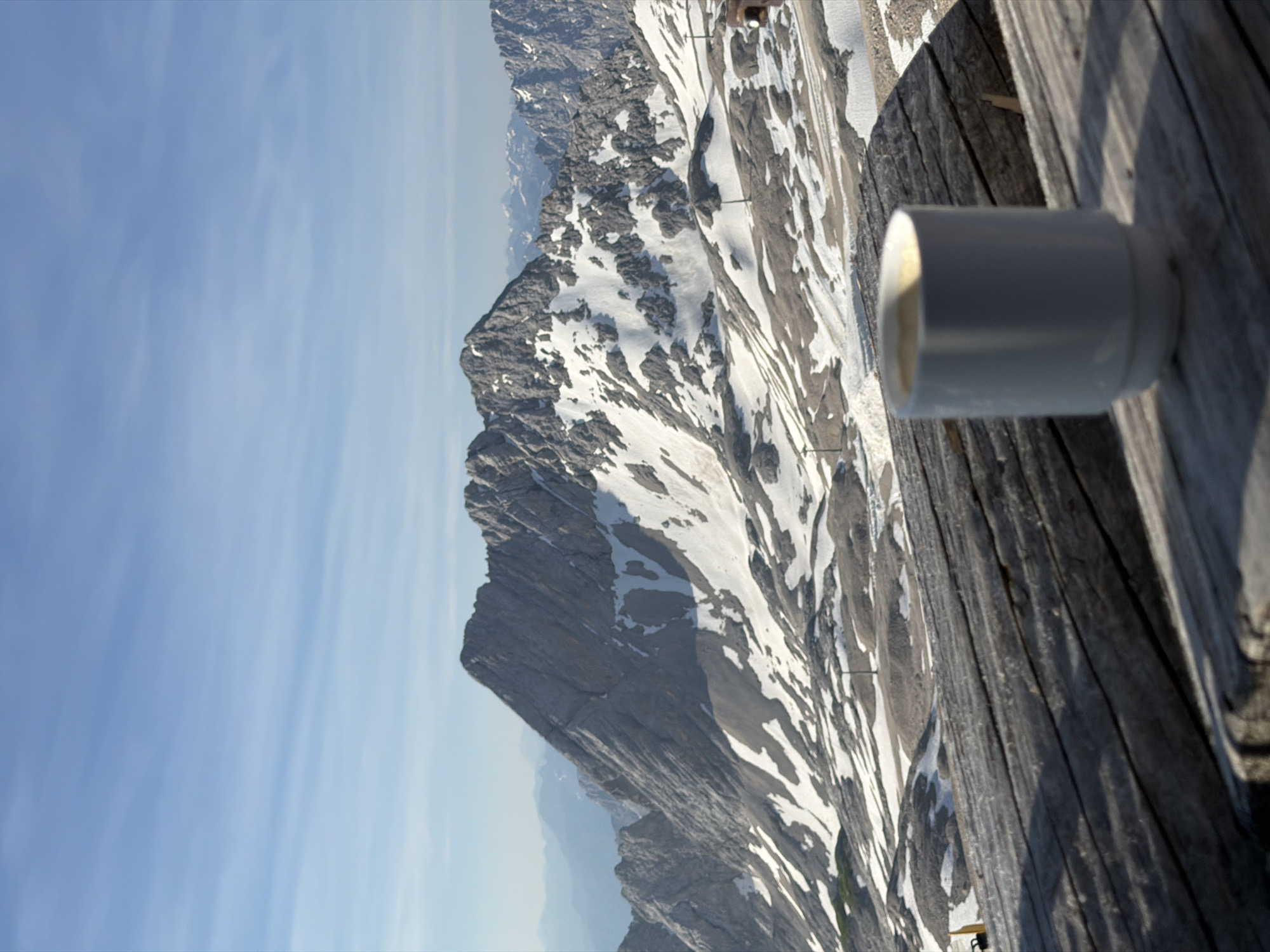The image depicts a scenic mountain landscape with a cup of coffee in the foreground. The mountains are rocky and partially covered in snow, suggesting a high-altitude location. The sky is clear and blue with some wispy clouds. In the foreground, a white ceramic mug filled with a light-colored beverage, likely coffee, sits on a weathered wooden surface. The wooden surface appears to be part of a table or bench, possibly on an outdoor viewing platform. The overall scene conveys a sense of tranquility and appreciation for nature.
This view is characteristic of the Zugspitze, Germany's highest peak, located near Garmisch-Partenkirchen. The landscape with its distinctive rocky peaks and snowfields is consistent with the alpine environment of the Zugspitze region. The presence of a coffee cup on what could be a terrace of a mountain hut or restaurant further supports this interpretation.