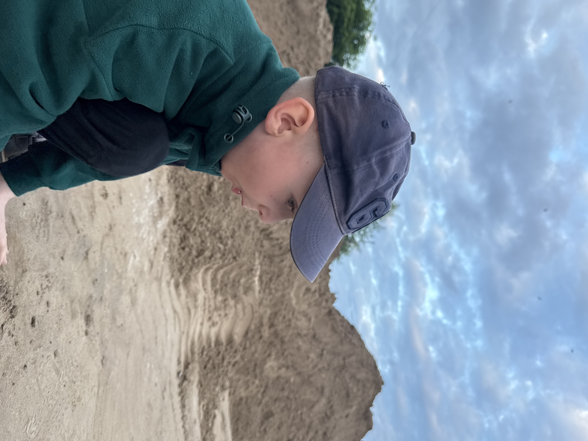 A young boy wearing a dark blue baseball cap and a dark green fleece jacket is crouching on sandy ground. He is looking to the right, with his mouth slightly open. In the background, there is a large pile of sand, possibly from construction or landscaping. The sky above is cloudy and overcast. The scene appears to be outdoors in a somewhat industrial or undeveloped area. The location is Chaikovsky, Russia.