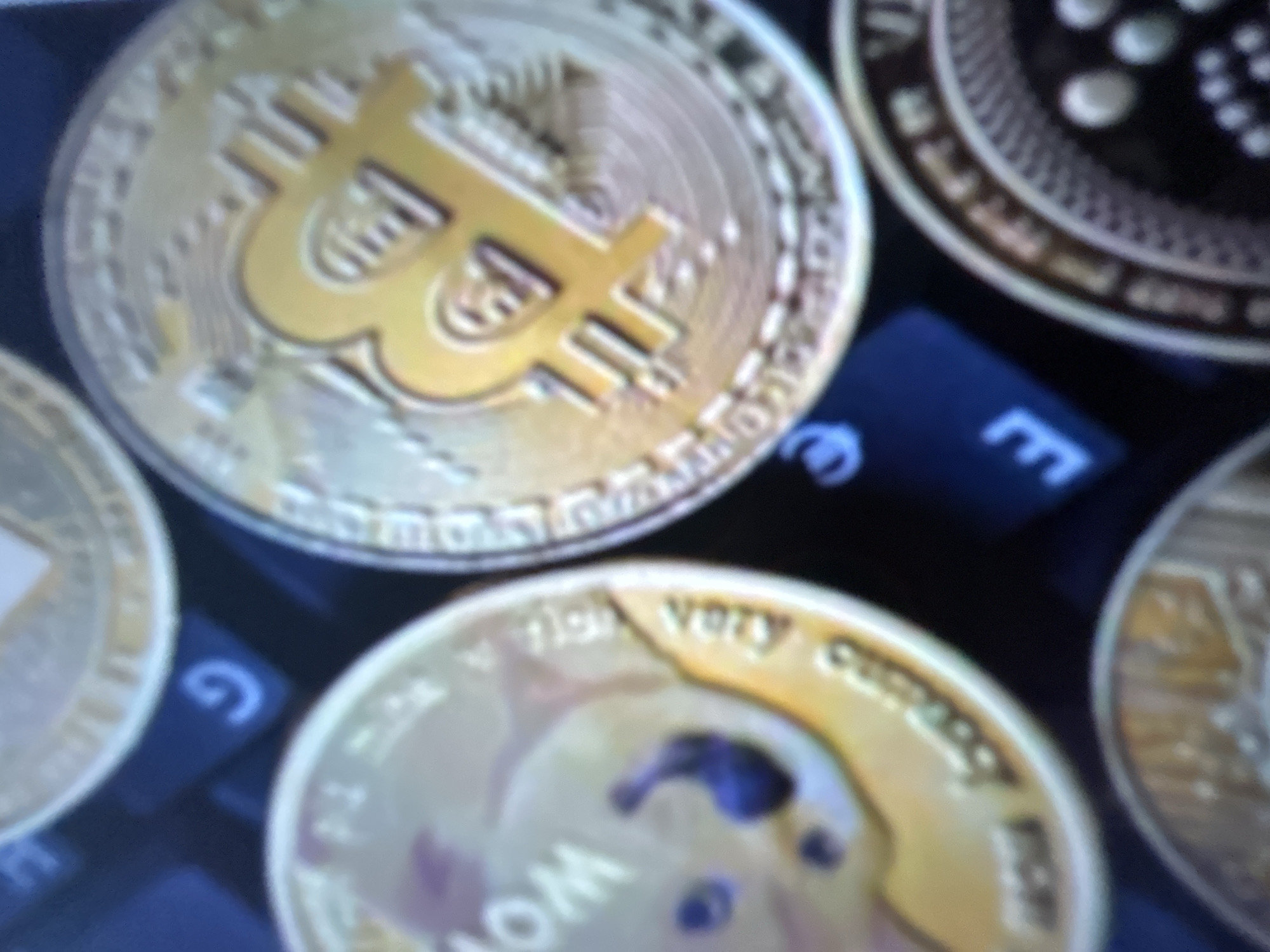 The image displays a collection of cryptocurrency coins, likely a physical representation of digital assets. The primary subject is a Bitcoin coin, prominently featured with its iconic "B" symbol. To its right, another coin is partially visible, with intricate circular patterns and text. Below the Bitcoin, a coin with the Shiba Inu dog image, characteristic of Dogecoin, is also visible. The coins appear to be laid on a dark surface, possibly a keyboard, as suggested by the presence of what looks like keys with letters such as "E" and "G" and a symbol resembling the Euro sign. The lighting is somewhat dim, and the focus is soft, giving the image a slightly blurred, ethereal quality. The arrangement of the coins suggests a display or a collection. There is no indication of people, a specific time of day, weather, or strong emotions. The setting appears to be indoors, with the background being a dark, non-descript surface. Visible text includes the "B" symbol on the Bitcoin, and partial inscriptions on the other coins, but they are not entirely legible due to the image quality. The mention of Gelendzhik, Russia, as a location context does not provide any visual cues within the image itself.