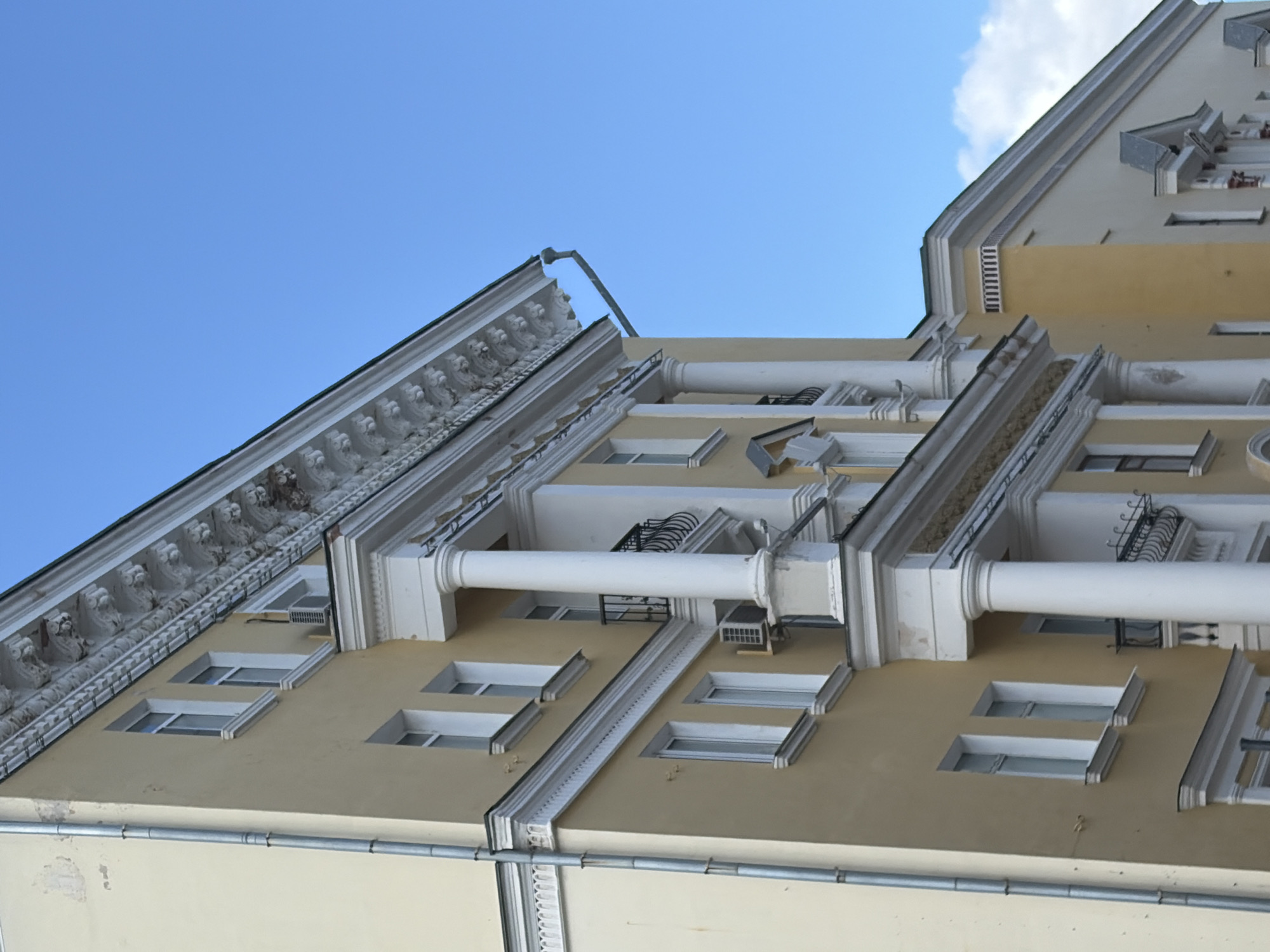 The image shows a close-up, low-angle shot of a pale yellow building with white architectural details, against a clear blue sky. The building features multiple windows with white frames and sills, some of which are divided into two panes. A prominent white column supports an overhang with decorative carvings. A metal downspout runs vertically along the left side of the building. There is a balcony with black wrought-iron railings. An air conditioning unit is visible mounted on the wall below a window. The building appears to be a residential or administrative structure, common in urban environments. The architectural style suggests it is likely from the mid to late 20th century. The scene is static, with no people or activity visible. The image does not provide sufficient information to definitively confirm its location in Nizhny Novgorod, Russia, but the architectural style is consistent with buildings found in Russian cities.
