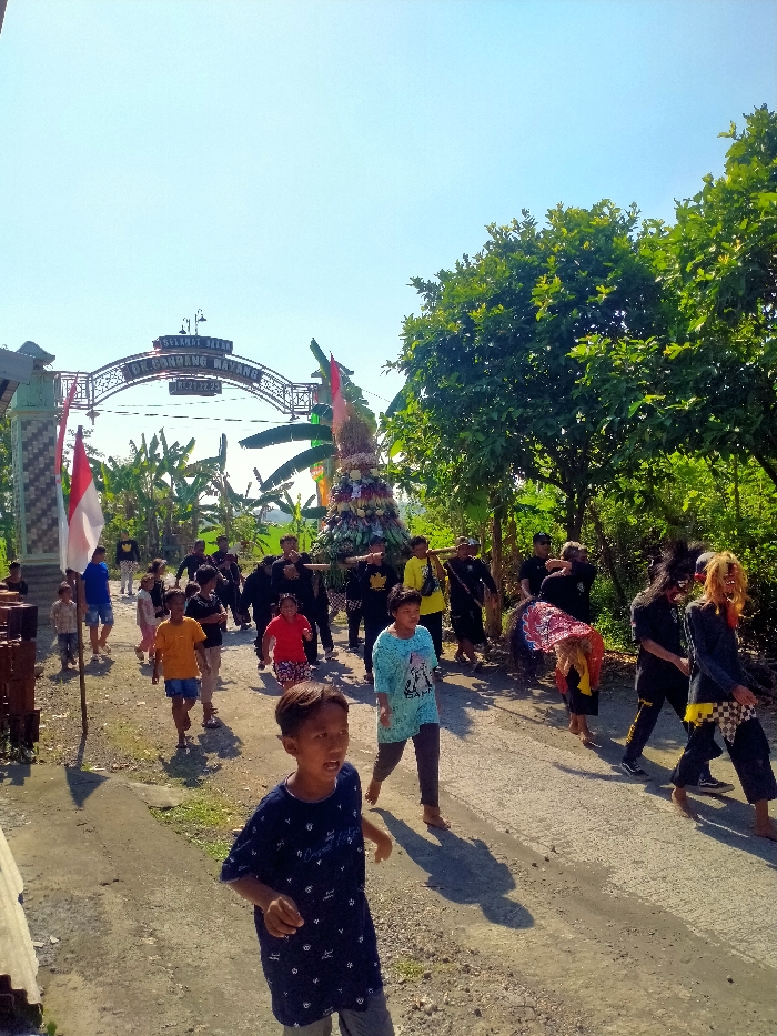 An outdoor daytime scene captures a cultural procession in Dukuh Gondang Wayang, Tanon, Indonesia. A group of approximately 20-25 individuals moves along an unpaved road under a clear blue sky, with bright sunlight casting distinct shadows. In the background, a metal archway marks an entrance, displaying the text "SELAMAT DATANG DUKUH GONDANG WAYANG RT.27 RW.9."

The central element of the procession is a large, conical offering structure, or *gunungan*, composed of fruits, vegetables, and rice stalks. This structure is carried on a wooden palanquin by at least eight men, primarily dressed in black shirts. Accompanying the *gunungan* are various participants, including two individuals on the right side wearing elaborate masks or face paint, one of whom also carries a dark, possibly animal-shaped, headpiece. Other participants, including children and adults in casual attire, walk alongside and in the periphery of the main group. In the far left background, an Indonesian flag (red and white) is affixed to a pole near a decorative pillar. Lush green trees and banana plants line the path, particularly on the right side and behind the archway, indicating a vegetated environment.