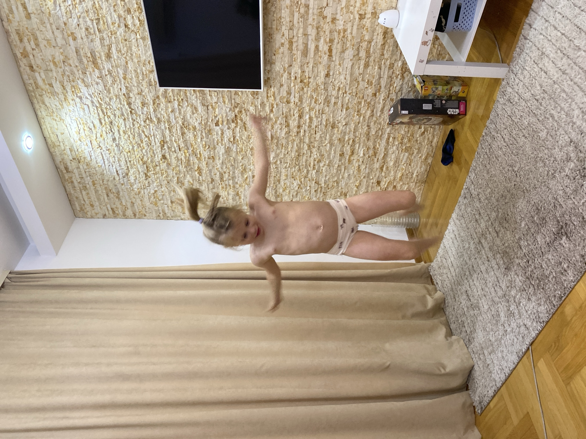 The image captures a young child, likely a girl with light-colored hair tied in a ponytail, mid-air, appearing to jump or fall playfully indoors. She is wearing light-colored underwear (briefs) and has her arms and legs splayed out, with a joyful or excited expression on her face.
The scene is an interior room. One prominent wall is covered with light-colored, stacked stone-like tiles, serving as a decorative feature. A black flat-screen television is mounted on this tiled wall. Below the television, to the right, is a white multi-shelf unit with a light blue basket on one shelf and a small white decorative item on top. Beneath this unit, on the wooden parquet floor, are several stacked boxes, one clearly labeled "STAR WARS" and another appearing to be "LEGO." A dark blue item of clothing is also on the floor.
The floor on the right side of the room is a wooden parquet, partially covered by a light gray, shaggy rug. On the left side of the room, beige or light brown curtains hang, extending towards the floor. A white ceiling or upper wall area is visible in the upper left, featuring a circular recessed light fixture that is illuminated. The overall impression is a brightly lit indoor space.