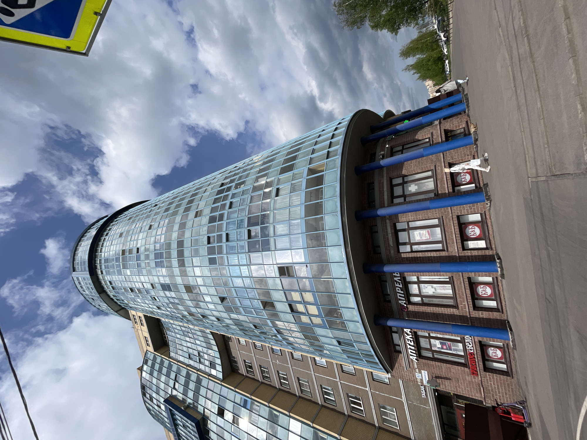 The image shows a cylindrical building in Saint Petersburg, Russia. The building has a rounded, glass-paneled facade and a brick base with blue cylindrical pillars. There are signs for a pharmacy ("АПТЕКА") and a liquor store ("Красное Белое"). A pedestrian can be seen walking on the sidewalk in front of the building. The sky is cloudy with patches of blue. A sign is also visible on the upper left.