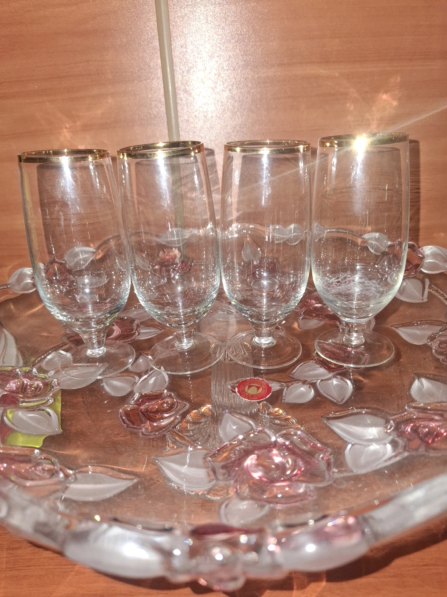 Four clear glass flutes with gold rims are arranged on a glass tray decorated with raised pink and clear rose and leaf motifs. The flutes are empty and stand upright on the tray, which has a slightly raised edge. A red and gold circular sticker is affixed to the tray near the base of the second flute from the left. The background appears to be a wooden surface, possibly a table or wall, with a vertical beige object running down the center. The lighting creates reflections and highlights on the glass surfaces and the background. This image was captured in Almaty, Kazakhstan.