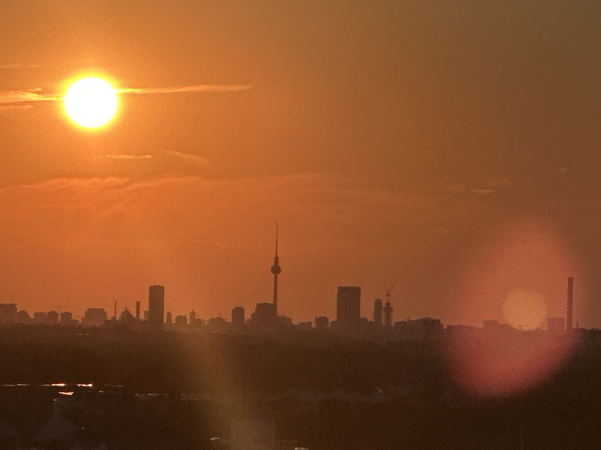 This is a photograph of the Berlin skyline at sunset. The sun is a bright, glowing orb in the upper left quadrant of the image, casting a warm orange and yellow hue across the sky. A silhouette of the Fernsehturm, Berlin's television tower, is visible in the center of the frame, rising above a cluster of other buildings. Other indistinct building shapes and construction cranes are also discernible against the dusky sky. The foreground is composed of a dark, silhouetted landscape, likely trees or rolling hills, with hints of reflected light on what appears to be water. The overall atmosphere is one of warmth and tranquility, typical of a city sunset.