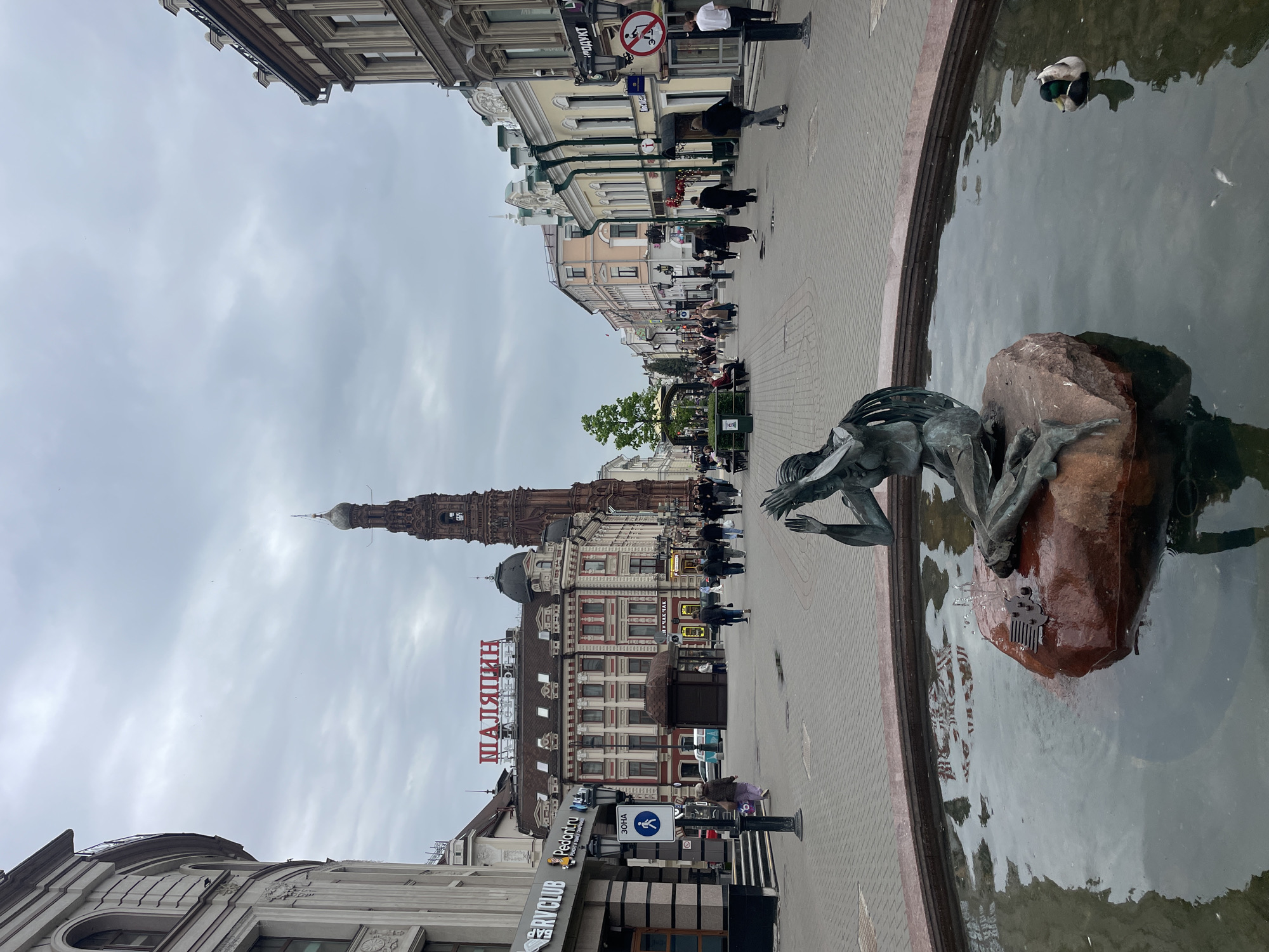 The image is a street scene in Kazan, Russia. The foreground features a fountain with a bronze statue of a boy perched on a rock. Two ducks are swimming in the fountain. A brick-paved pedestrian area stretches into the distance, flanked by buildings of varying heights and architectural styles. The "Shalyapin" building is visible. The Suyumbike Tower is visible in the background. People are walking along the street. The sky is overcast.