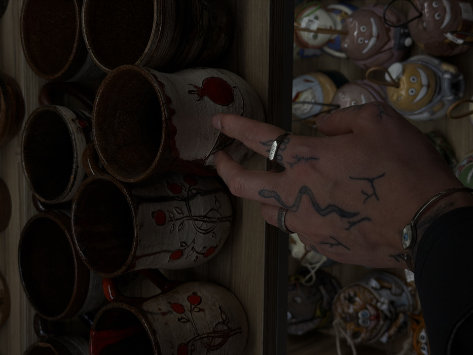 A close-up view depicts a person's tattooed right hand reaching forward to touch a handmade ceramic mug displayed on a wooden shelf. The hand features a prominent snake tattoo winding across its back and forearm, along with several smaller, abstract stick-figure-like tattoos on the fingers and back of the hand. A textured silver band ring is on the index finger, and a plainer silver band is on the ring finger. A silver bracelet with an "evil eye" charm is visible on the wrist, emerging from a dark sleeve.
The wooden shelf holds numerous artisanal ceramic mugs, predominantly light-colored (off-white/cream) with dark brown rims and interiors. Many are decorated with traditional Armenian motifs, including stylized red pomegranates and branched designs, some with red accents or handles. The hand is interacting with a mug featuring a large red pomegranate. Below, a second wooden shelf displays an assortment of smaller, brightly colored ceramic figurines, including whimsical animal or character shapes. The scene is consistent with a craft stall or shop within the Vernissage market in Yerevan, Armenia, known for its extensive collection of local handicrafts.