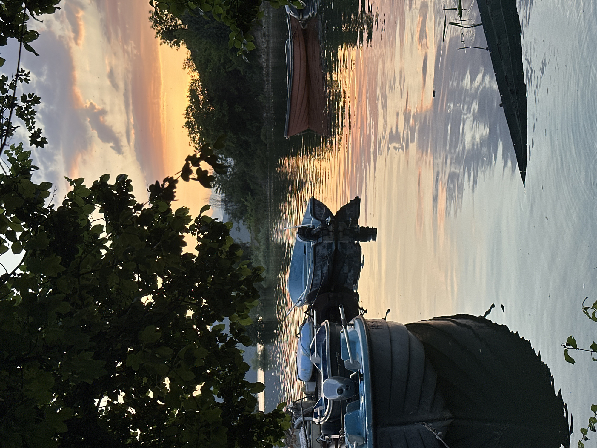 The image depicts a serene sunset scene over a body of water, likely a lake or river, in Glavatartsi, Bulgaria. The sky, painted with soft gradients of orange, gold, and light blue, features scattered clouds that catch the warm light of the setting sun. This vibrant sky is beautifully reflected on the calm surface of the water, creating gentle, rippled patterns that mirror the colors above.
In the foreground and mid-ground, several small boats are visible. Most prominent is a light blue boat with an outboard motor, positioned towards the left. Another dark-hulled boat is partially seen on the far right edge of the frame. Further out on the water, a distinct brown boat floats near the distant shoreline, which is densely covered with trees and bushes, appearing as a dark silhouette against the brighter sky and water. Lush, dark green foliage from trees and bushes frames the left side of the composition, partially obscuring the view of the boats and water.
No people or direct activity are visible, contributing to the tranquil and peaceful atmosphere. The scene captures a quiet moment at dusk, characterized by soft light and the stillness of the water. There is no visible text in the image.