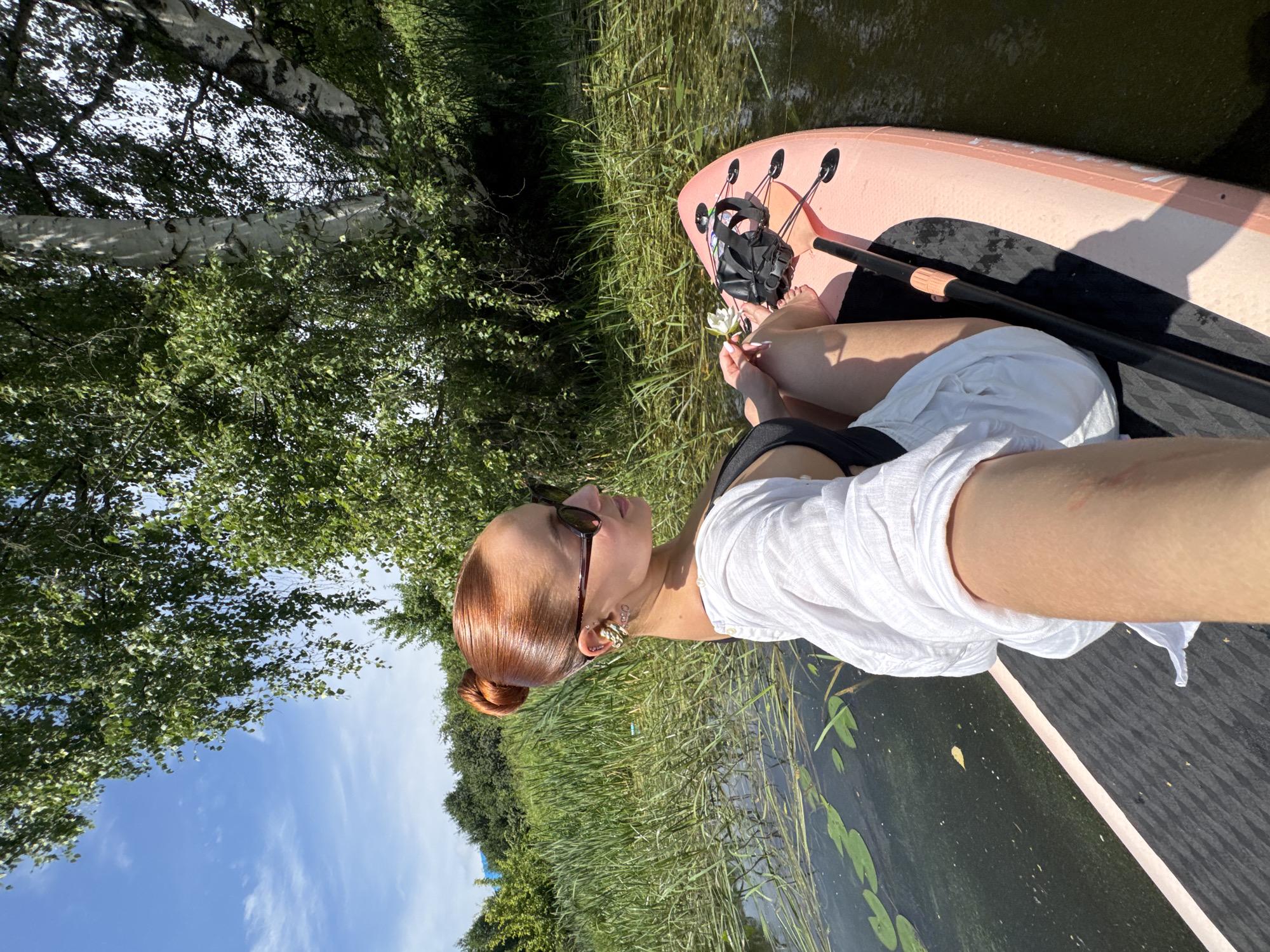 A woman with red hair pulled back in a bun is sitting on a paddleboard in a body of water, holding a white flower. She is wearing a black swimsuit and a white cover-up, and sunglasses. The paddleboard is pink and black. The water is surrounded by tall green reeds and trees. The sky is blue with white clouds. The scene appears to be in a natural, outdoor setting, possibly a lake or river, which may be located in the city of Chastyye, Russia.