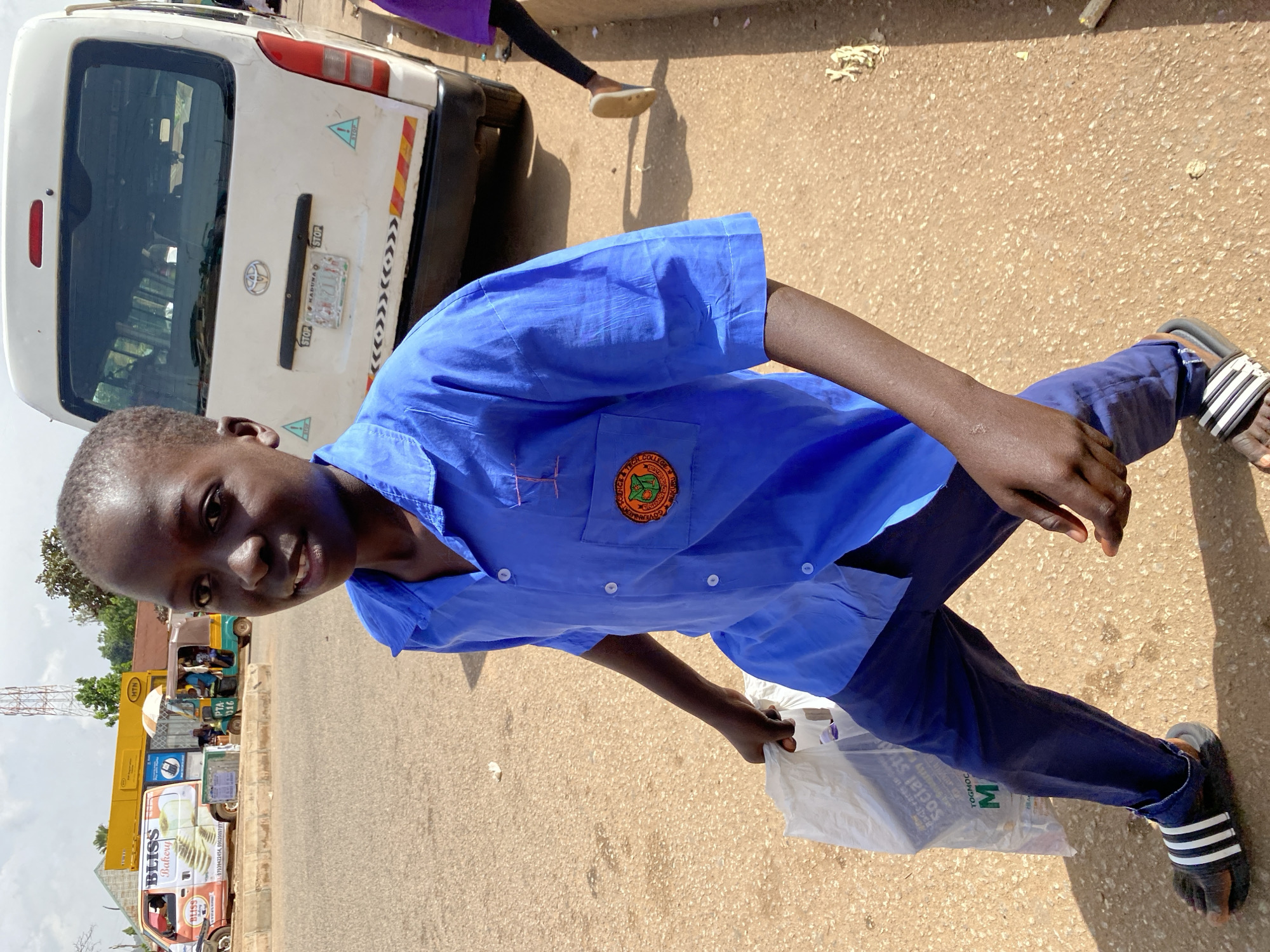 A young boy with dark skin, wearing a blue collared shirt and navy pants, walks on a dirt road in Bukuru, Nigeria. He is carrying a white plastic bag in his left hand. The boy is looking towards the camera with a neutral expression. In the background, a white van is parked on the side of the road, and a yellow building with "MTN" signage is visible. To the left of the van, a smaller vehicle with an umbrella shade is present, with people inside. The environment appears to be a street with a few trees and telephone poles. The lighting suggests it is daytime.