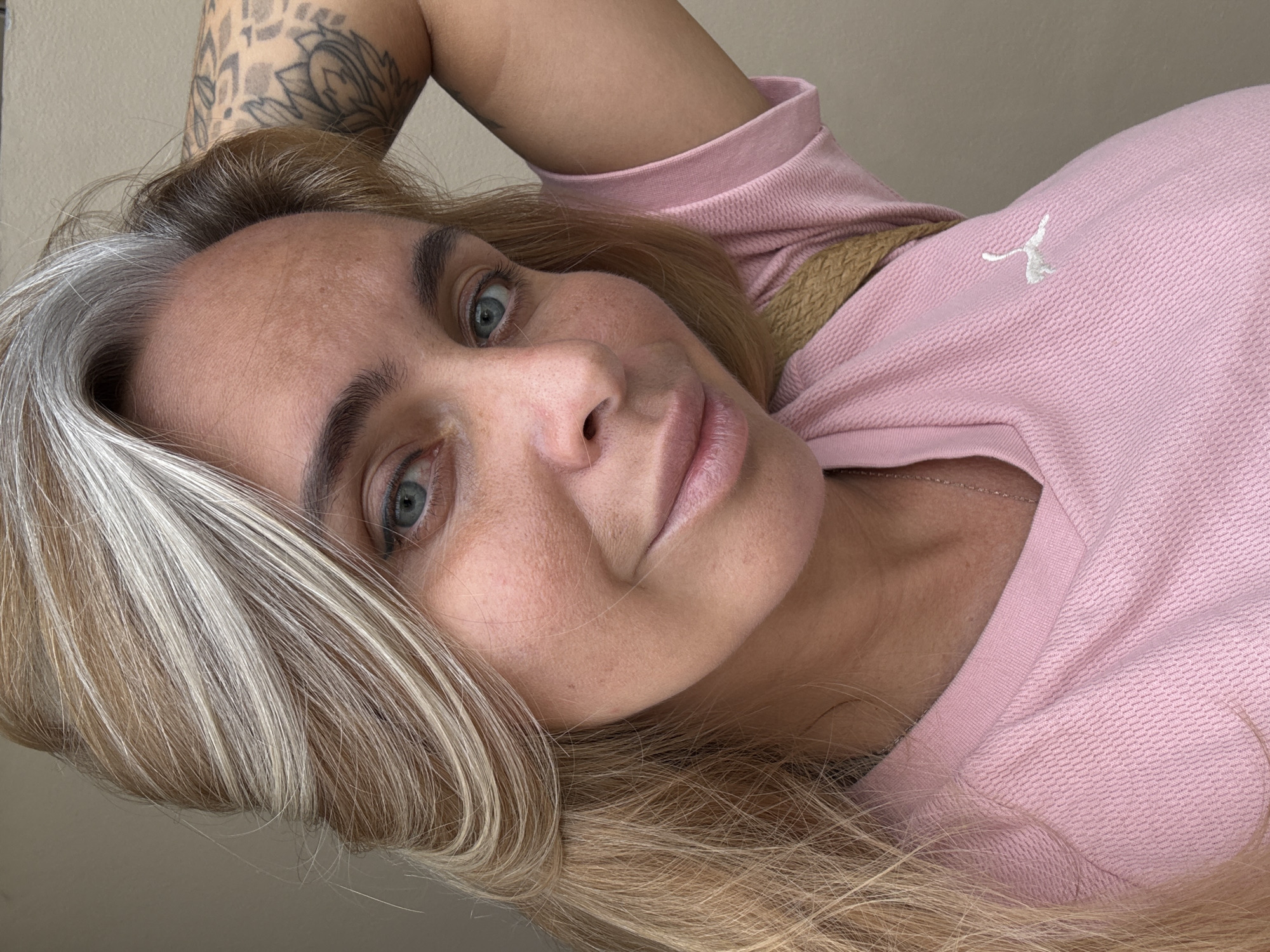 A close-up, selfie-style shot features a woman with blonde and gray hair, blue eyes, and fair skin. She is wearing a pink t-shirt with a white Puma logo. Her left arm is bent, with her hand resting on her head, revealing a tattoo on her upper arm. The background is a neutral, plain wall. The image was captured in Ko Samui District, Thailand.