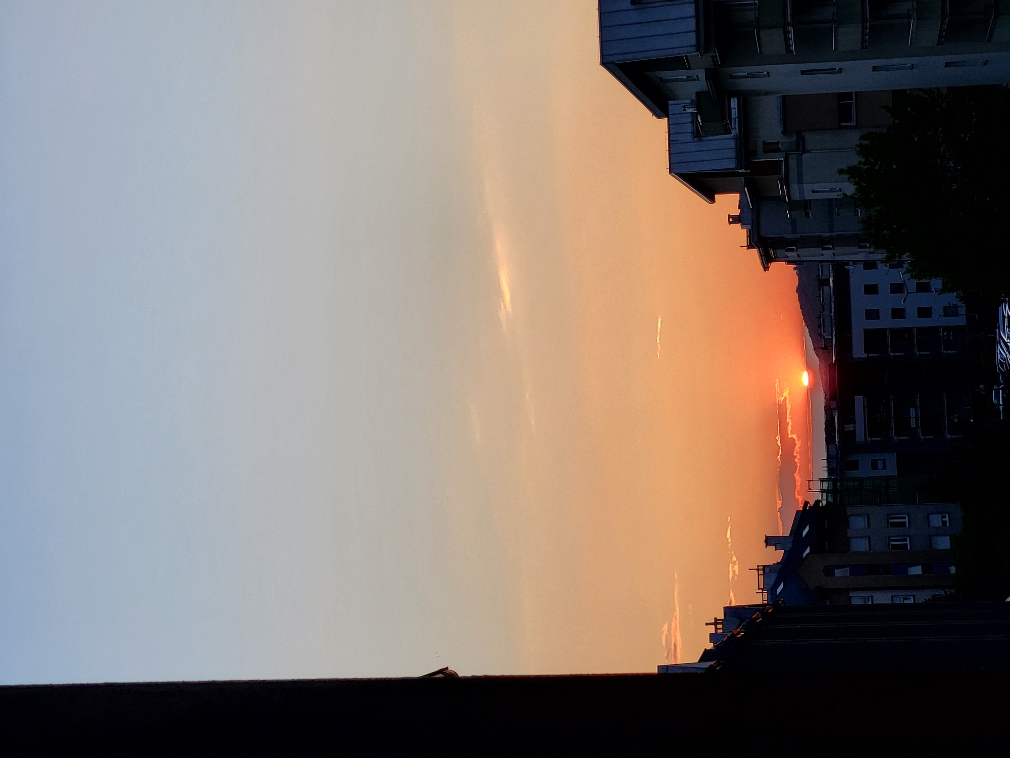 The image depicts a sunset in an urban environment, likely in Unknown, Unknown. The sky transitions from a pale blue on the left to a warm orange hue near the horizon, where the sun is setting. The sun's light is partially obscured by clouds or haze near the horizon. Buildings line the right side of the image, appearing silhouetted against the bright sunset. The tops of more buildings are also visible in the lower right portion of the image, with a dark barrier at the bottom of the frame possibly being a rooftop or balcony edge.