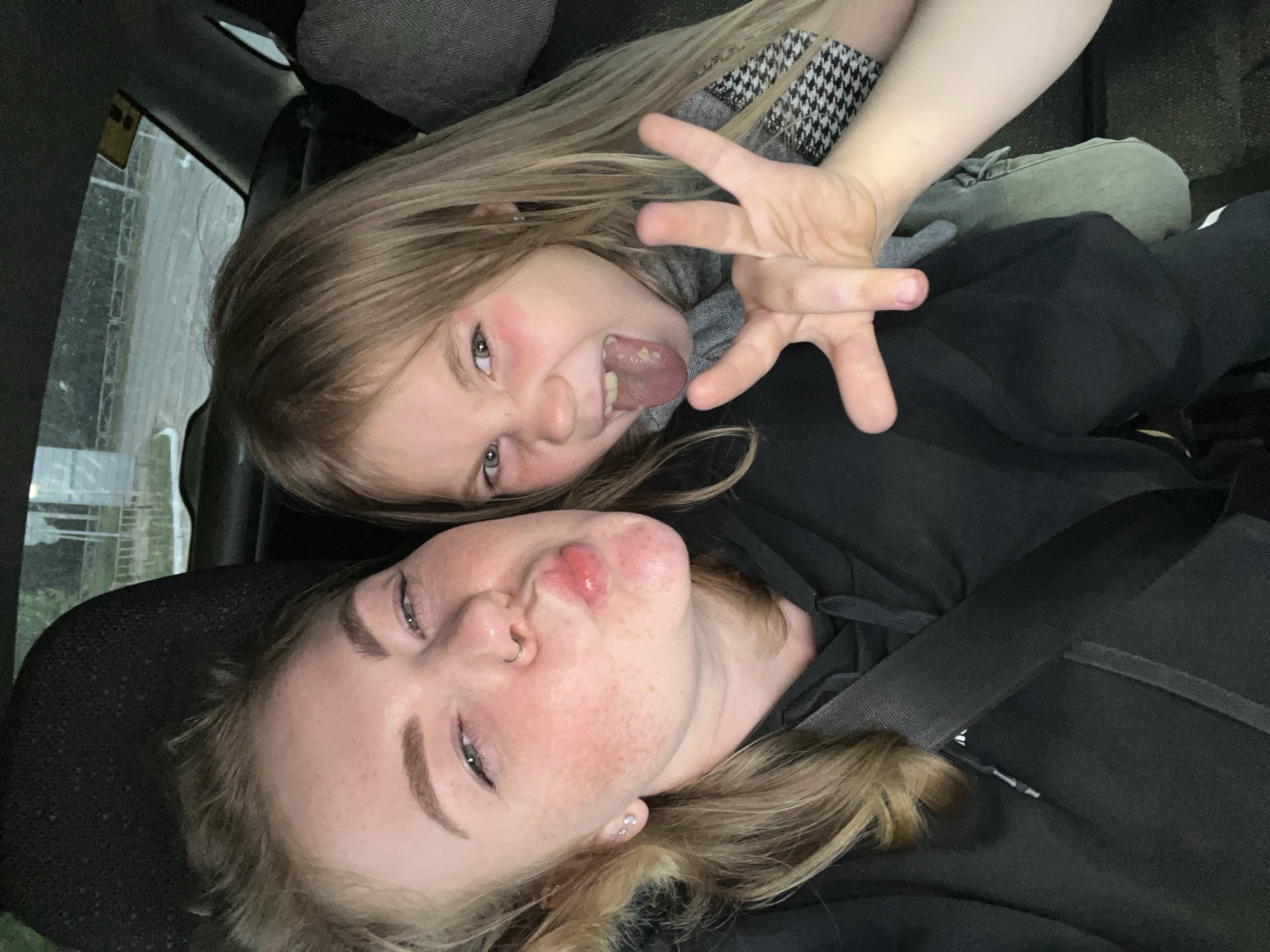 Two people are inside a car, in what appears to be Chaikovsky, Russia, based on the car's interior and the background view. A young woman in the foreground is making a kissing face, her lips pursed. She has freckles on her face, a nose ring, and is wearing a black hoodie with a seatbelt across her chest. To her right, a young girl is sticking her tongue out playfully, making a hand gesture with her fingers spread. She has rosy cheeks and is wearing a grey scarf with a black and white pattern. The background shows a street scene with trees and a fence, suggesting they are parked or driving in a city or town.