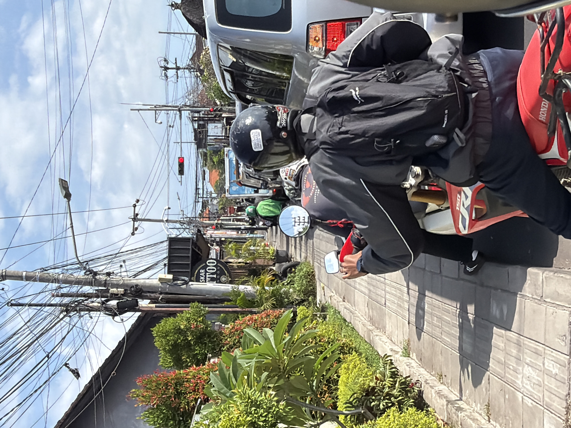 The image shows a street scene in North Kuta, Indonesia. A person wearing a helmet, jacket, and backpack is sitting on a motorcycle, with a silver car in front. Several other motorcycles and vehicles are visible further down the road. A "SKAR TATTOO" sign can be seen on a building, along with power lines above. The environment includes greenery and plants lining the street. A traffic light is showing red in the distance.