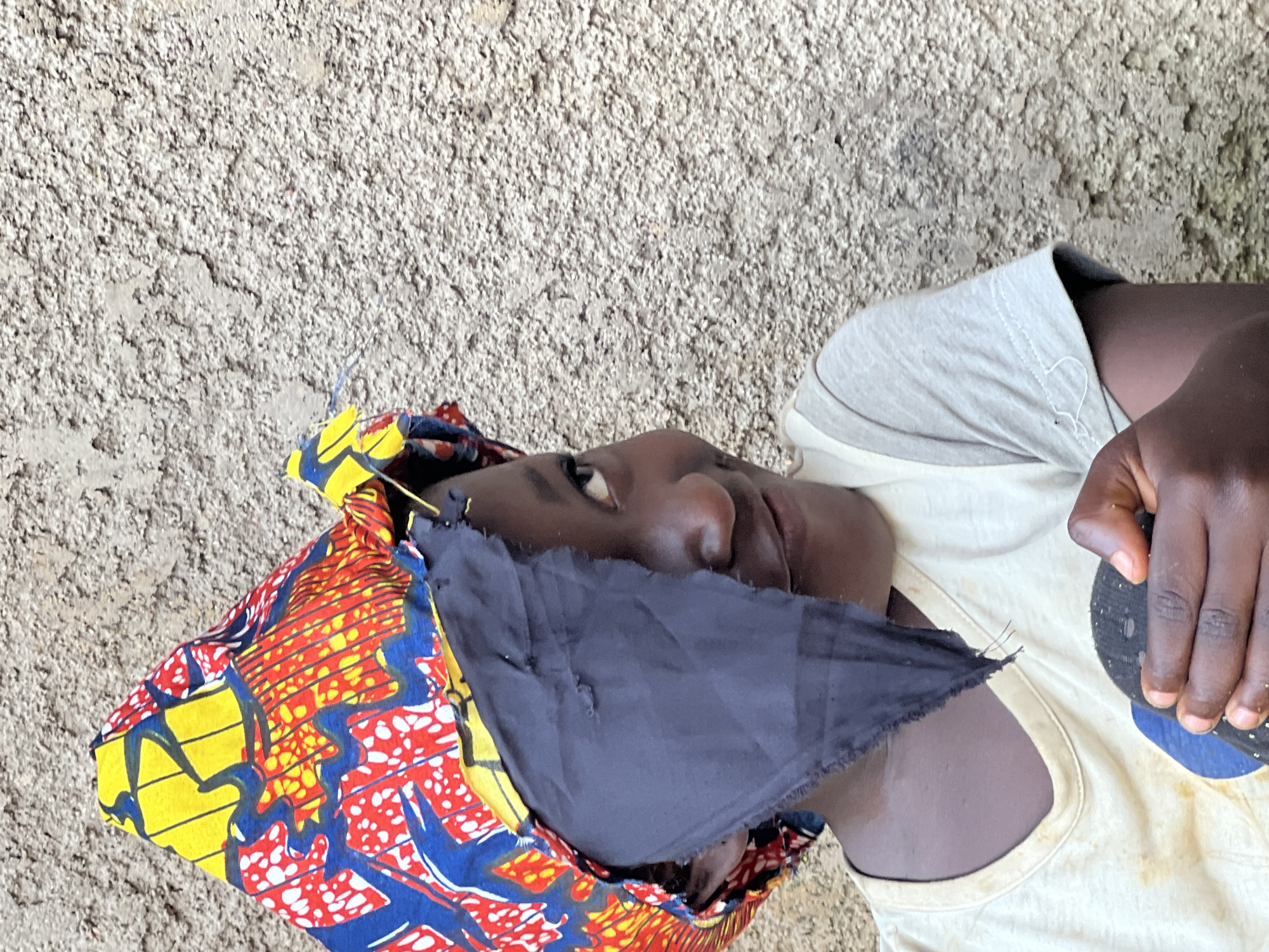 A young child with dark skin, wearing a colorful African print headscarf partially covering their face, stands in front of a rough, textured beige wall. The headscarf features vibrant patterns of red, yellow, and blue, with a piece of dark fabric obscuring the child's left eye. The child is looking to their right with a slight smile. They are wearing a light-colored t-shirt with a V-neck, which appears to have some stains. In their hands, they are holding a dark object, possibly a sock or a small toy. The environment suggests an outdoor setting, with the textured wall and possibly dirt on the ground visible. The overall impression is of a candid portrait of a child in a humble, outdoor environment, likely in a residential area within the city of Jos, Nigeria.