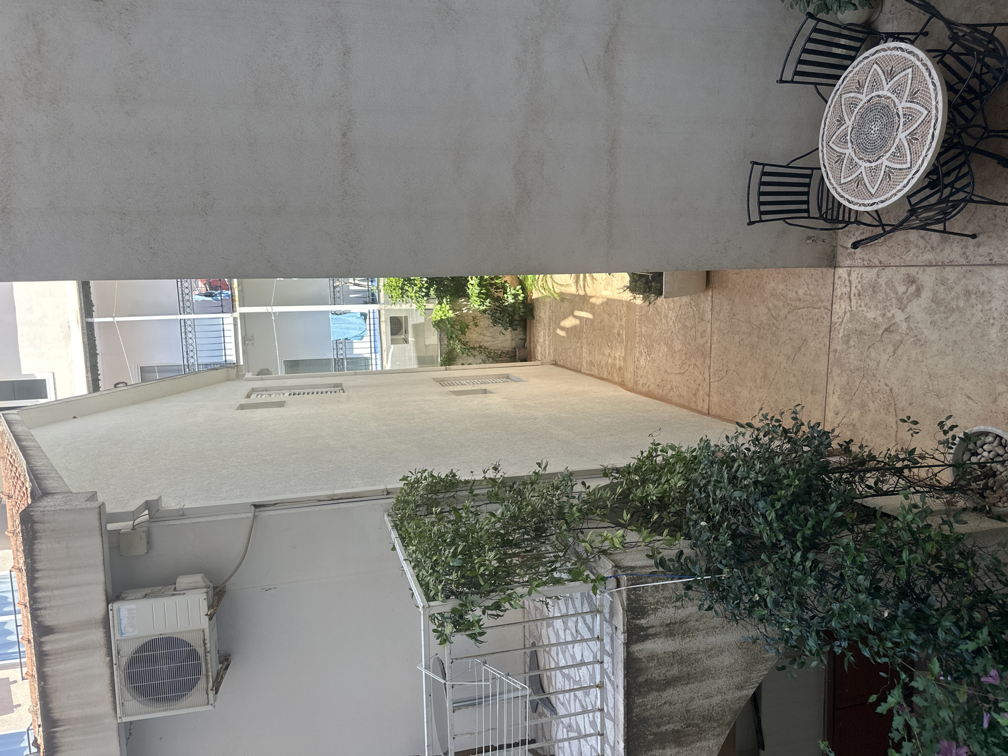 The image shows a narrow courtyard between two buildings in Budva, Montenegro. On the left, a white building has an air conditioning unit mounted on its exterior wall. A white metal railing with a patterned tiled floor is visible on a lower level, with lush green foliage climbing up the railing. A clothes drying rack is also present.
The building on the right is a lighter shade of gray and has a textured surface. In the foreground of this building's side, a black metal bistro set with a circular mosaic-tiled table and four chairs is arranged on a patterned concrete floor.
The far end of the courtyard opens up to another building with multiple balconies, some adorned with laundry hanging out to dry. Potted plants are scattered throughout the courtyard, and shadows suggest the sun is high in the sky. The overall scene depicts a typical urban courtyard, likely residential, in a European coastal town.