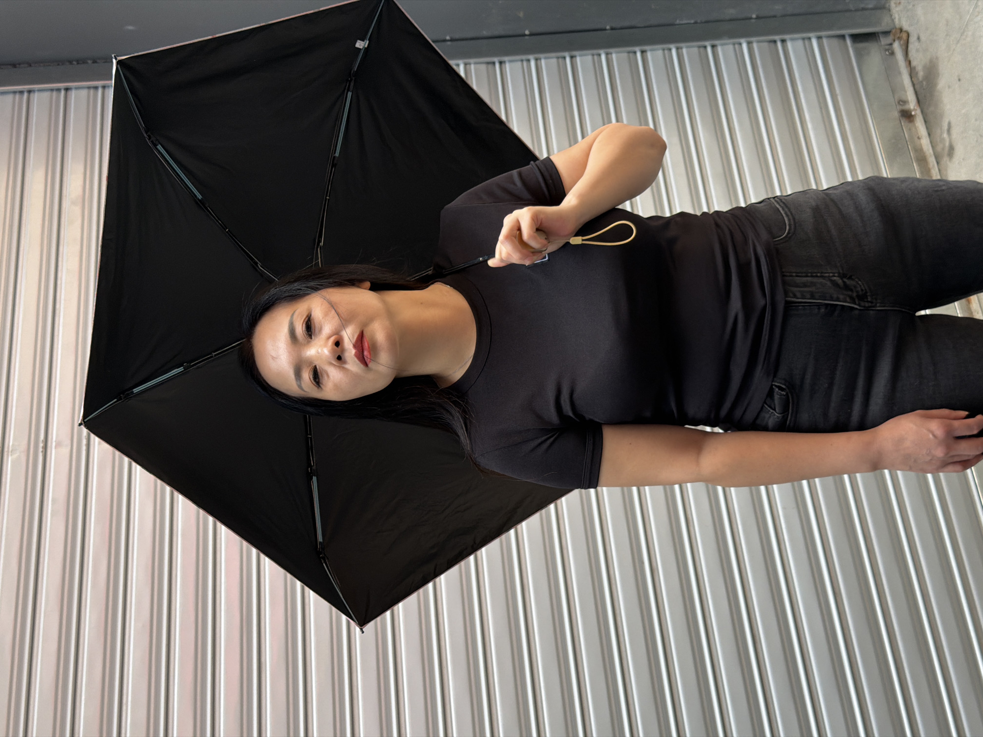 A medium shot captures an Asian woman standing outside, holding a black umbrella. The woman is looking directly at the camera with a neutral expression. She is wearing a black t-shirt and dark jeans. The background is a metallic, corrugated roll-up door. The scene appears to be in an urban setting.