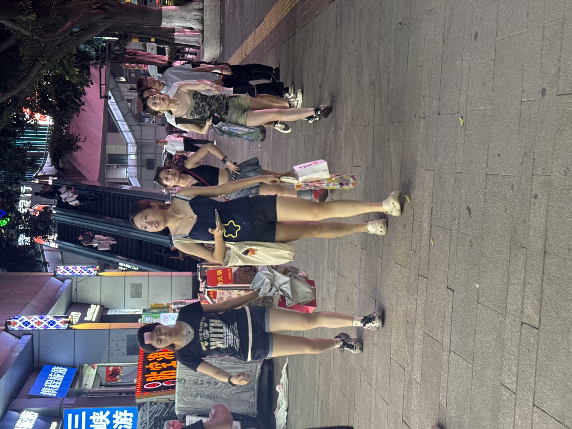 The image depicts a group of people walking on a pedestrian walkway in Yuzhong District, China, at night. The walkway is paved with stone tiles and illuminated by neon signs and building lights. In the background, an escalator is visible, with people ascending and descending. Several individuals are walking towards the camera, including three young women in casual attire. The woman in the foreground on the left is wearing a black t-shirt with a graphic print and denim shorts, with a pink hair roller on her forehead. The woman next to her is wearing a black tank top and a denim skirt. The third woman is wearing a camouflage-patterned tank top and shorts. Other people are walking in the background, and some are looking at their phones. The scene suggests a lively urban environment with commercial activity.