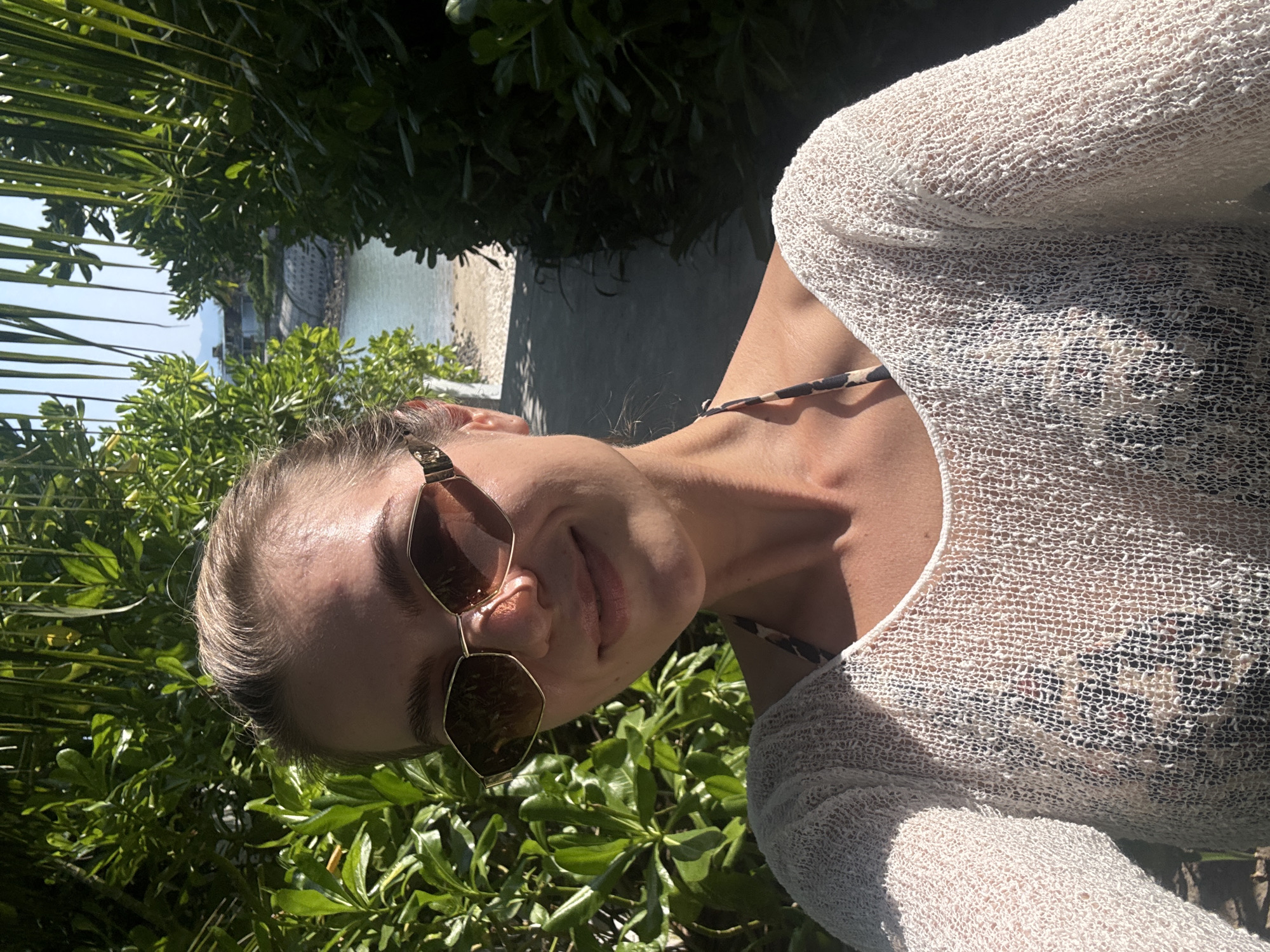The image shows a woman wearing sunglasses and a white cover-up over a patterned swimsuit. She is positioned outdoors, surrounded by green foliage. In the background, there is a glimpse of a beach and ocean. The location is unknown.