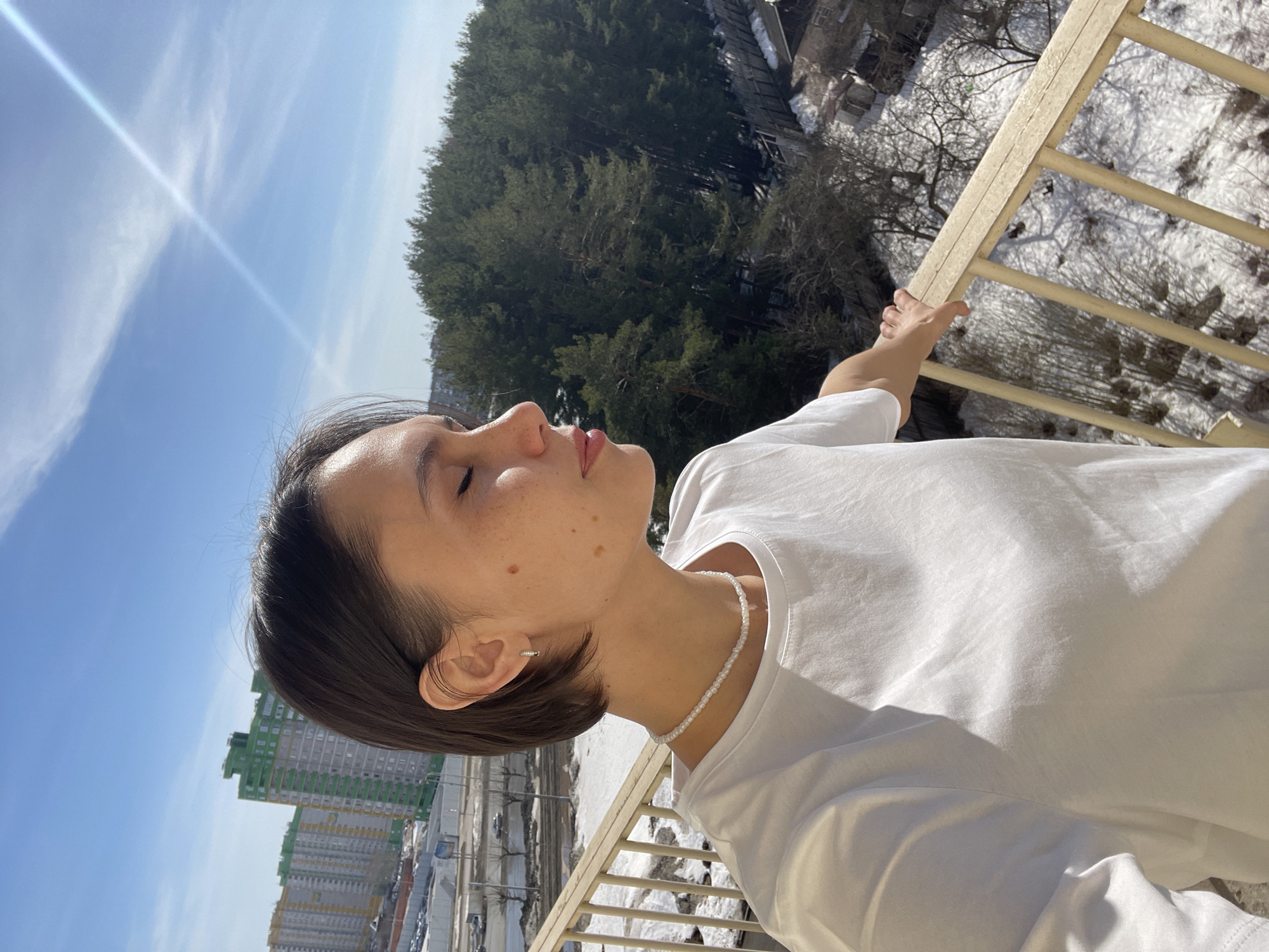 A woman with short, dark hair, fair skin, and wearing a white t-shirt and pearl necklace is standing on a balcony, holding the railing with her right hand. Her eyes are closed and her face is turned upward, seemingly enjoying the sun. The balcony overlooks a cityscape with tall residential buildings in the distance and a snowy, wooded area below. The location is Izhevsk, Russia, based on visual cues consistent with architectural styles and urban planning found in that city. The sky is blue with some cloud cover.