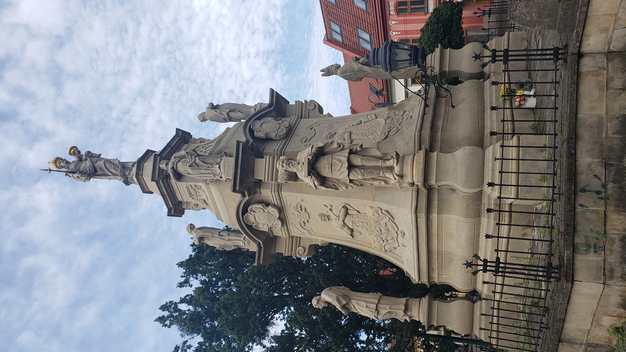 This is a low-angle, full shot of the Holy Trinity Column located in the center of the main square ( námestie) in Prešov, Slovakia. The column is a large, ornate Baroque monument made of light-colored stone, with multiple tiers and statues. At the very top, a statue of the Virgin Mary holding the Christ Child is visible, wearing a golden crown and holding a scepter. Below her are several other statues of saints and religious figures adorning the various levels of the column. The monument is surrounded by a black, ornate metal fence. In the background, a large green tree stands to the left, and buildings with red-tiled roofs are visible on the right. The sky is partly cloudy with a bright, overcast day. There are no people visible in the immediate vicinity of the monument, though there is a lamppost and some flowers in a bucket on the ground near the base of the column.