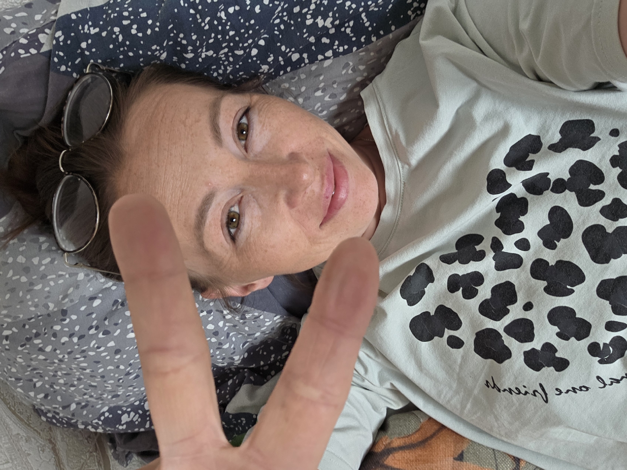 A woman is lying on a bed, possibly in Unknown, Unknown. She has brown hair and eyes. She is wearing glasses and a light-colored shirt with black spots. Her right hand is raised, displaying a "peace" or "victory" sign. The bed has a gray and blue patterned comforter.
