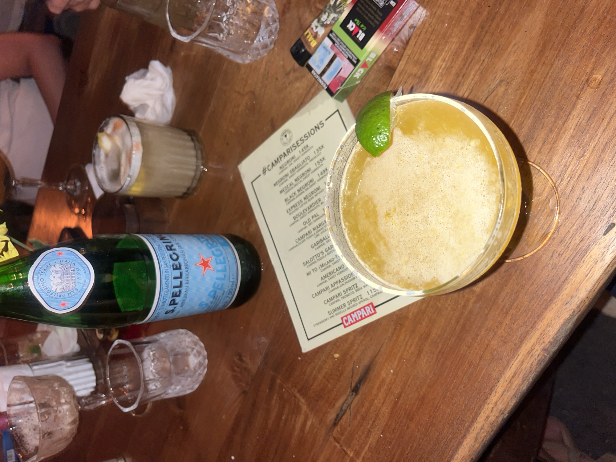 The image depicts a wooden table setting, likely in a bar or restaurant. A lime-garnished cocktail occupies the right side of the frame. Next to the cocktail is a cocktail menu advertising "#CAMPARISESSIONS" and listing various Campari-based drinks with associated prices. A bottle of San Pellegrino water is placed near the left side of the menu. Further left, there are partially filled and empty glasses. A pack of Black cigarettes can be seen in the upper center of the frame. The location is in Kuta Selatan, Indonesia, based on the menu language.