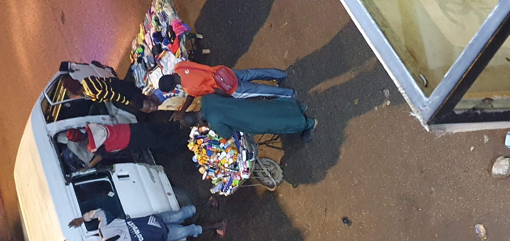 In Bukuru, Nigeria, a white mini-van with its doors open is parked on a paved road. Several individuals are gathered around the vehicle and a nearby stall. One person, wearing a white long-sleeved shirt with "COWBOYS" printed on it, stands by the open driver's side door, gesturing with their left hand. Inside the van, two people are partially visible. To the right of the van, a person in a red hoodie is standing with their back to the camera, while another person in a green robe is leaning over a wheelbarrow filled with various items, including numerous small bottles and containers. A table laden with folded clothing is visible further to the right. The ground appears to be a mixture of asphalt and loose gravel. Shadows are cast on the ground, suggesting it is daytime.