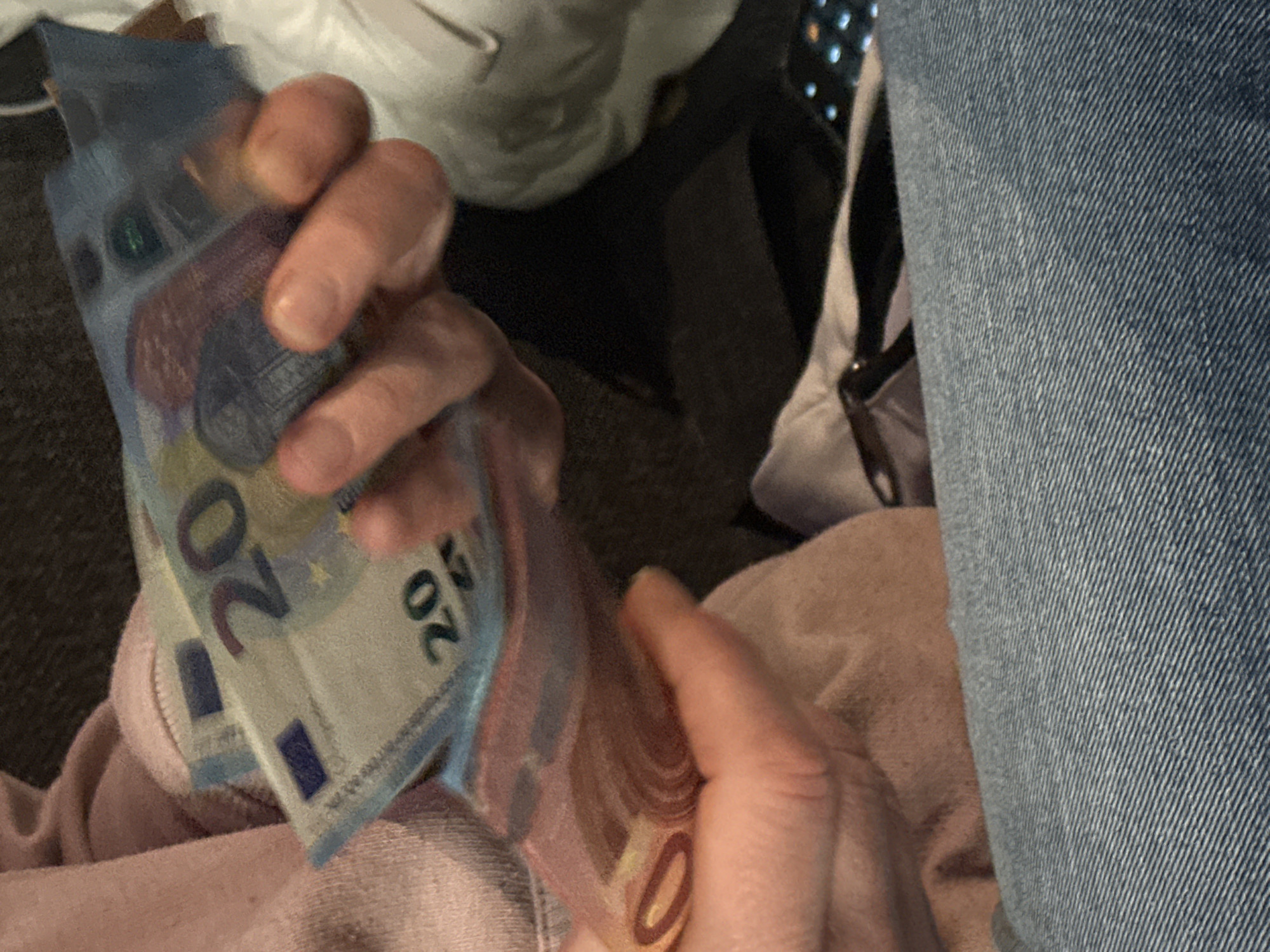 A person holds a stack of 20 Euro bills in their hand. The person is wearing a light pink sweatshirt. The person is sitting next to a person wearing jeans. There is a white object and a black object in the background. The specific location in Varna, Bulgaria, is not determinable from the image.