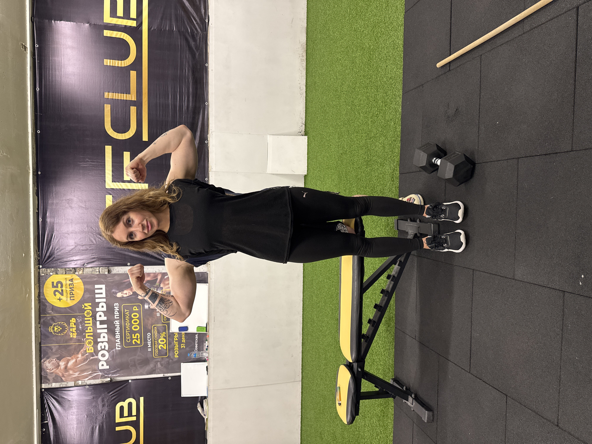 A woman stands posing in what appears to be a gym or fitness club, flexing both biceps and looking towards the camera with a slight smile. She has long, wavy brown hair and is wearing a black short-sleeved top, black leggings, and black and white athletic shoes. Tattoos are visible on her left forearm.
The background features a large black banner with gold lettering that reads "FCLUB" vertically, indicating the name of the establishment. To the woman's left, another large banner in black and gold promotes a "БОЛЬШОЙ РОЗЫГРЫШ" (Big Raffle/Giveaway). Visible text on this banner includes "ЦАРЬ" (Tsar/King), "+25 ПООЩРИТЕЛЬНЫХ ПРИЗА" (+25 incentive prizes), "ГЛАВНЫЙ ПРИЗ" (Main Prize) of a "СЕРТИФИКАТ 25 000 Р" (Certificate for 25,000 Russian Rubles), "II МЕСТО" (II Place) offering a "ГОДОВАЯ СКИДКА 20%" (Annual Discount 20%). The raffle is scheduled for "31 дека" (31 Dec), and a partial address "Советская," (Sovetskaya) is also visible.
The floor is a mix of black rubber gym tiles and green artificial turf. To the woman's right, a black and yellow adjustable workout bench is visible, along with a black dumbbell and a light-colored wooden stick or bar lying on the floor. The setting suggests a modern fitness environment. The woman appears confident and happy while showcasing her strength.