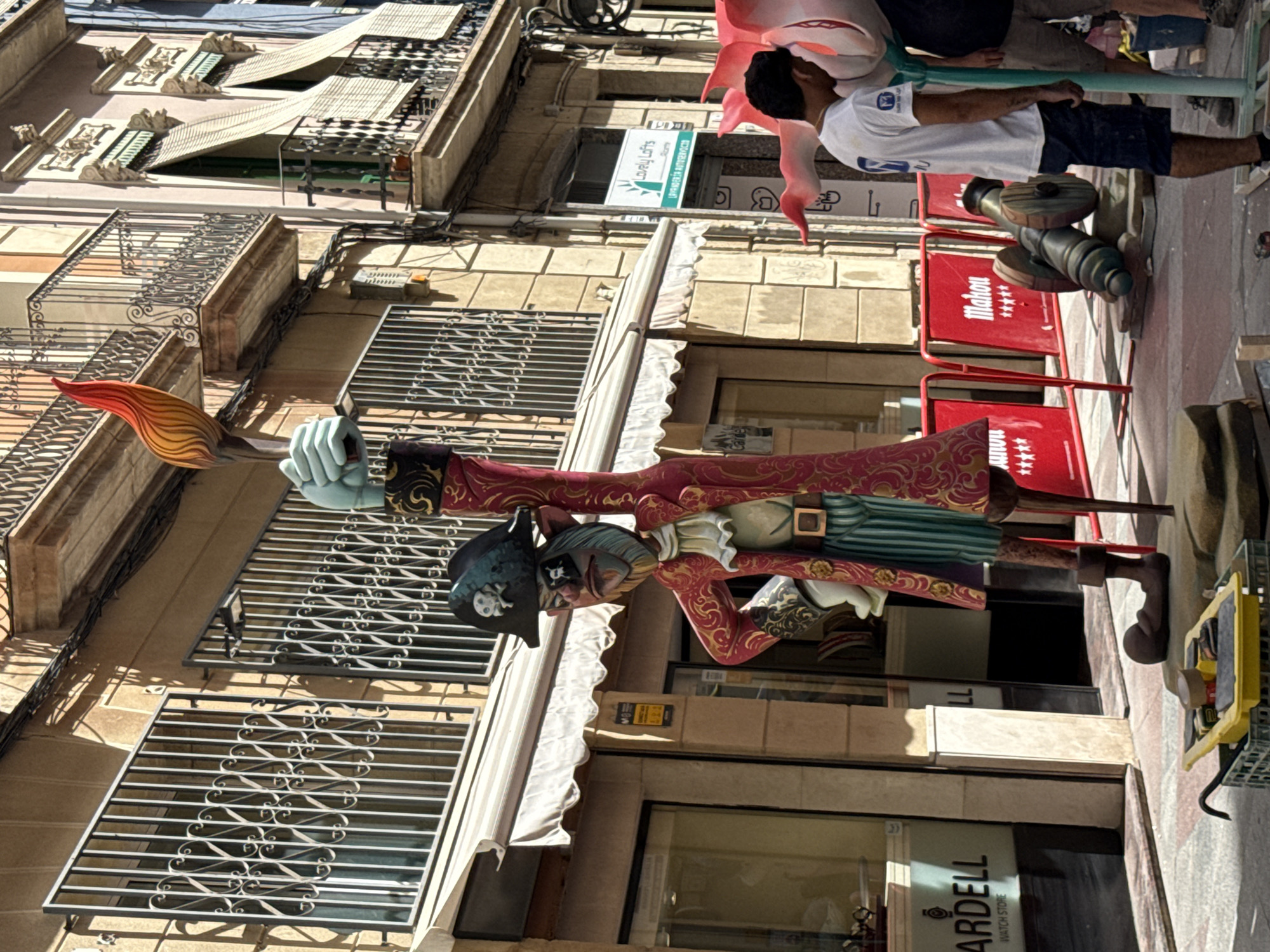 An outdoor, eye-level, medium shot captures a street scene with a large, colorful effigy of a pirate holding a torch. The effigy, dressed in a red coat with gold trim, striped pants, and a pirate hat with a skull emblem, stands prominently in the foreground. The pirate's left hand is raised, holding a flaming torch depicted with vibrant red and orange colors. The effigy appears to be part of a street display or festival.
In the background, buildings line the street, featuring balconies with ornate railings, windows with metal grates, and awnings. A shop window with the text "ARDELL WATCH STORE" is visible on the left. Red barriers with "Mahou" branding are placed along the street, and a cannon sculpture is situated behind them. Two men, one wearing a white t-shirt and shorts and the other a dark t-shirt and shorts, are visible to the right of the effigy, seemingly observing or interacting with the display. The ground is paved with cobblestones. The lighting suggests it is daytime, with shadows cast by the structures and the effigy.
