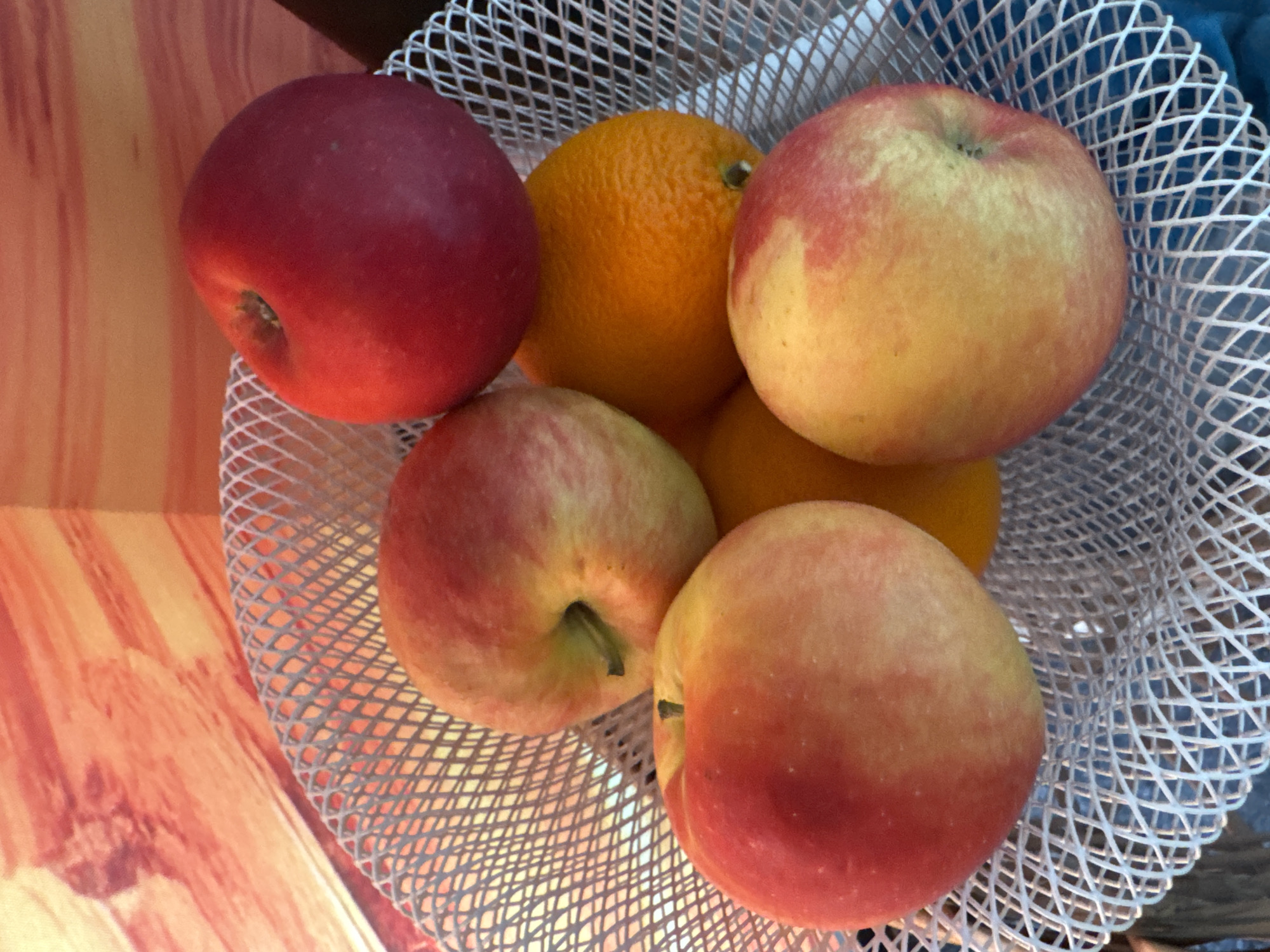 The image presents a close-up, high-angle view of a selection of fresh fruits contained within a white mesh basket. The subjects are six pieces of fruit: four apples and two oranges. The apples exhibit a gradient of colors, ranging from deep red to yellow-red, with some showing more pronounced red blush over a yellow base. One apple, positioned prominently on the left, is almost entirely red. The two oranges are bright orange and display the typical textured skin of citrus fruit.
The objects in the scene include the white, possibly plastic, mesh basket which holds the fruit. The basket's woven pattern is clearly visible. The fruits appear to be ripe and in good condition.
The setting appears to be indoors, with the basket resting on a surface that features a distinct pattern of reddish-brown and orange stripes, possibly a wooden table or a decorative mat. A darker, indistinct background, possibly a wall or curtain with a bluish tint, is visible behind the basket.
There is no discernible activity or event taking place; the image is a still life composition of fruit in a basket.
Notable details include the healthy appearance of the fruits and the clear texture of both the fruit skins and the mesh basket. The lighting is soft and even, suggesting an indoor environment without direct sunlight. There are no people visible, nor is there any visible text. The location context for this indoor scene is Boreti, Montenegro.