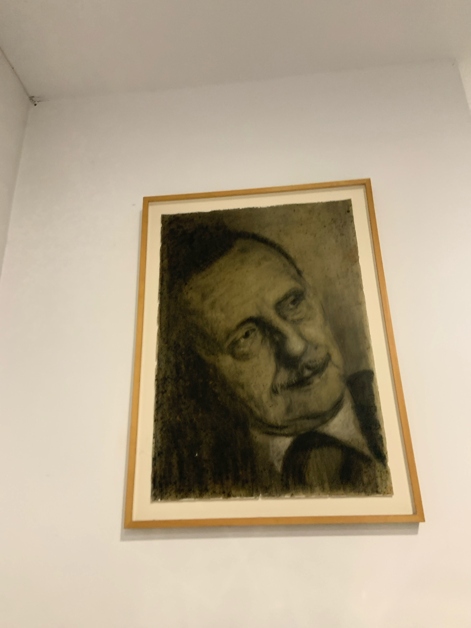 The image shows a framed charcoal drawing of a man's head and shoulders, hanging on a white wall. The drawing is in portrait orientation and is tilted slightly. The man in the drawing has dark hair, a mustache, and appears to be wearing a suit. The background of the drawing is dark and textured. The frame is a light wood color. The wall is plain white and appears to be a standard interior wall. The image does not contain any recognizable landmarks or features that would indicate a specific location in Alcorcón, Spain.
