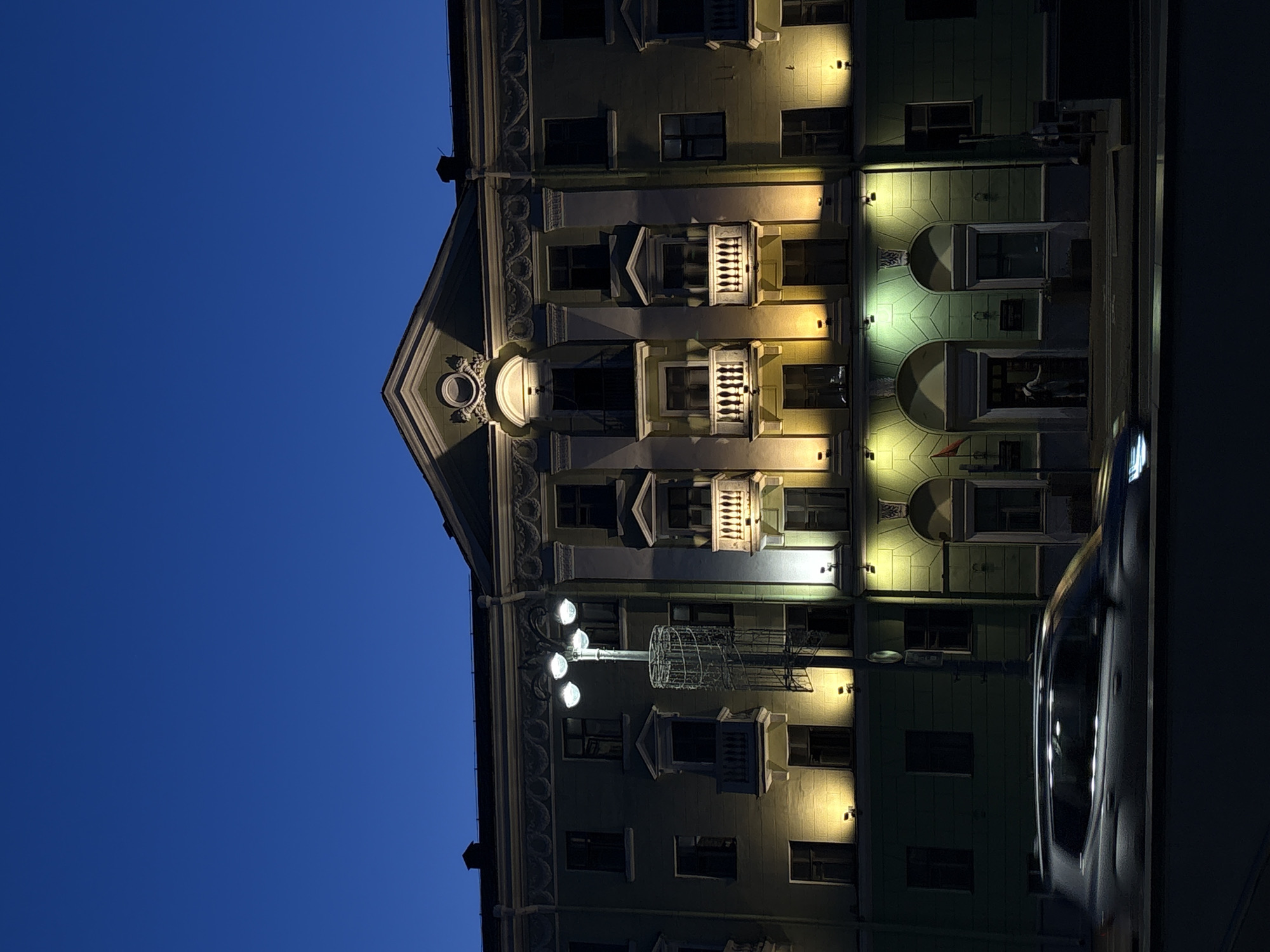 The image shows a building in Minsk, Belarus, at night. The building's facade is lit with warm-toned lights, accentuating architectural details like balconies, arched windows, and decorative moldings. A streetlamp stands in front of the building. The sky is a deep blue, contrasting with the illuminated building. A car is parked in front of the building, partly visible on the right.