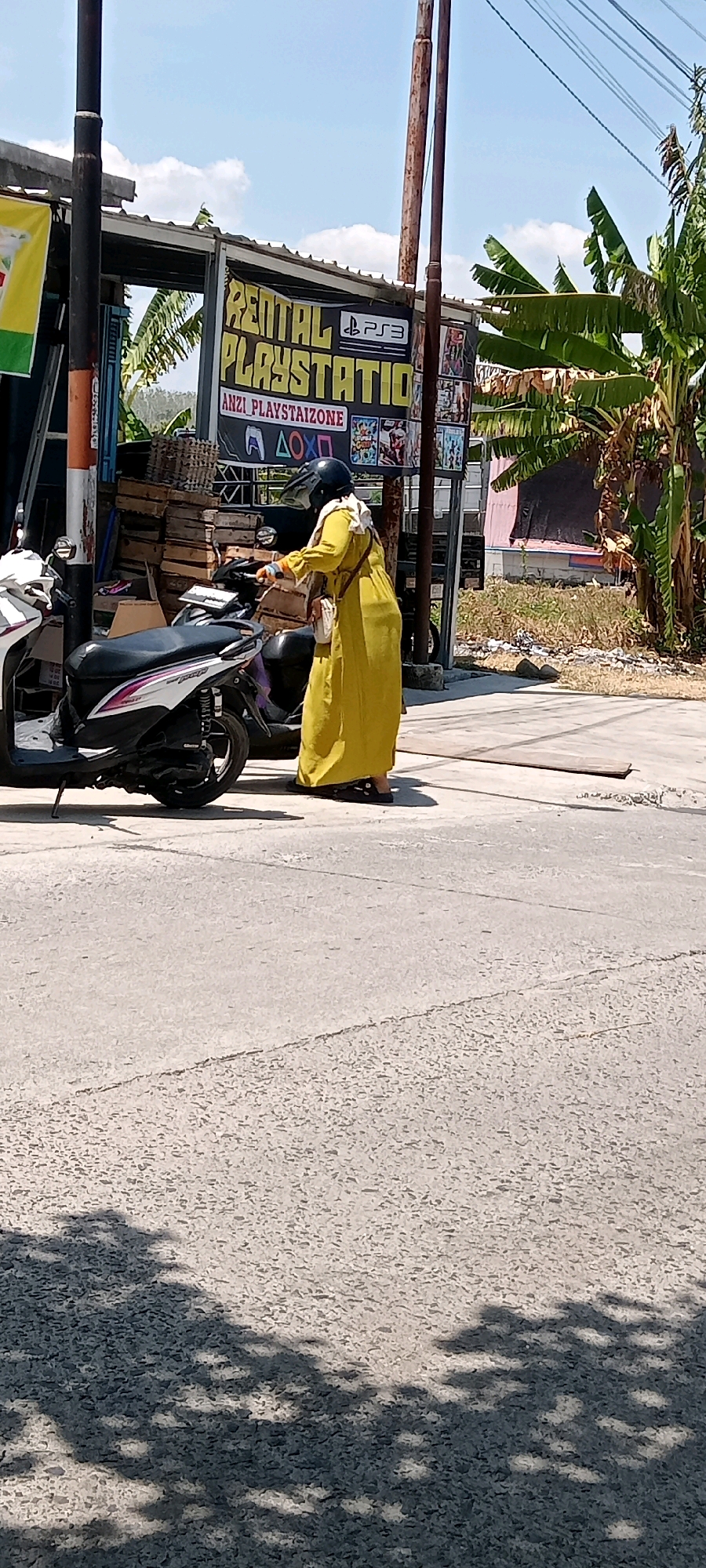 In Desa Karangmojo, Indonesia, a person wearing a yellow full-length dress and a black helmet stands next to a parked white scooter. The person appears to be interacting with a sign behind them, which reads "RENTAL PLAYSTATION" and "ANZI PLAYSTAIIZONE" with PlayStation symbols. The background features banana trees, a building with a corrugated metal roof, and stacked wooden crates. The scene is set on a paved road under a clear, bright sky.