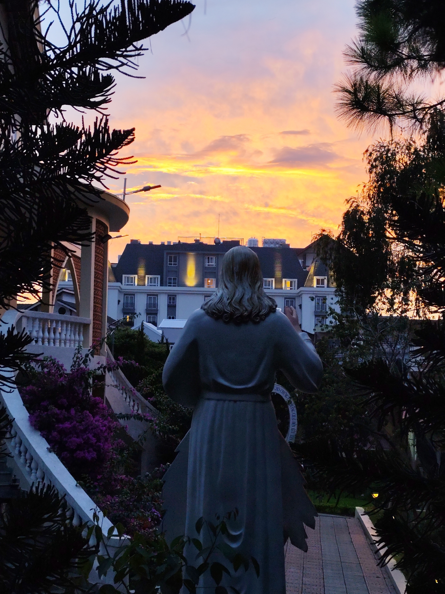 A low-angle shot captures a statue from the back, facing a building under a vibrant orange sunset sky in Da Lat, Vietnam. The statue, with long hair, is draped in a robe. The architecture is multistory, with white walls and dark rooflines, with lights visible in some windows. The foreground is partially framed by dark, silhouetted trees. To the left of the statue is a white balustrade with purple flowers. The statue stands on a brick pathway.