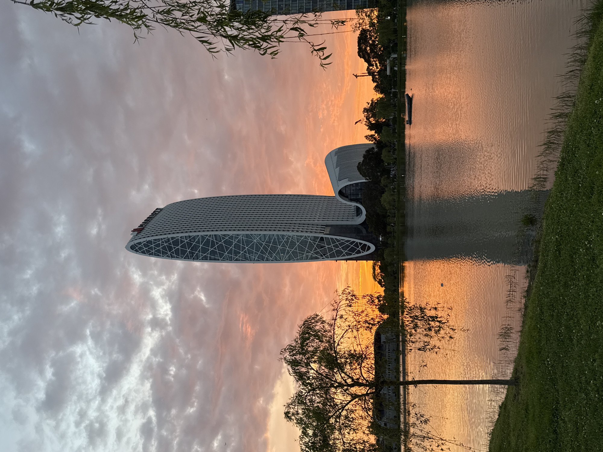 The photograph depicts a scenic view at sunset, showcasing a modern building with a unique, elongated oval shape and a patterned exterior. It is situated near a body of water, likely a lake or a river, where the building and the colorful sky are reflected. The sky displays a mix of orange, pink, and grey clouds. Some trees and vegetation are visible along the water's edge. There's also a glimpse of a boat or small structure on the water. Based on the architectural style and the landscape, this is likely the Batumi Technological University in Batumi, Georgia.