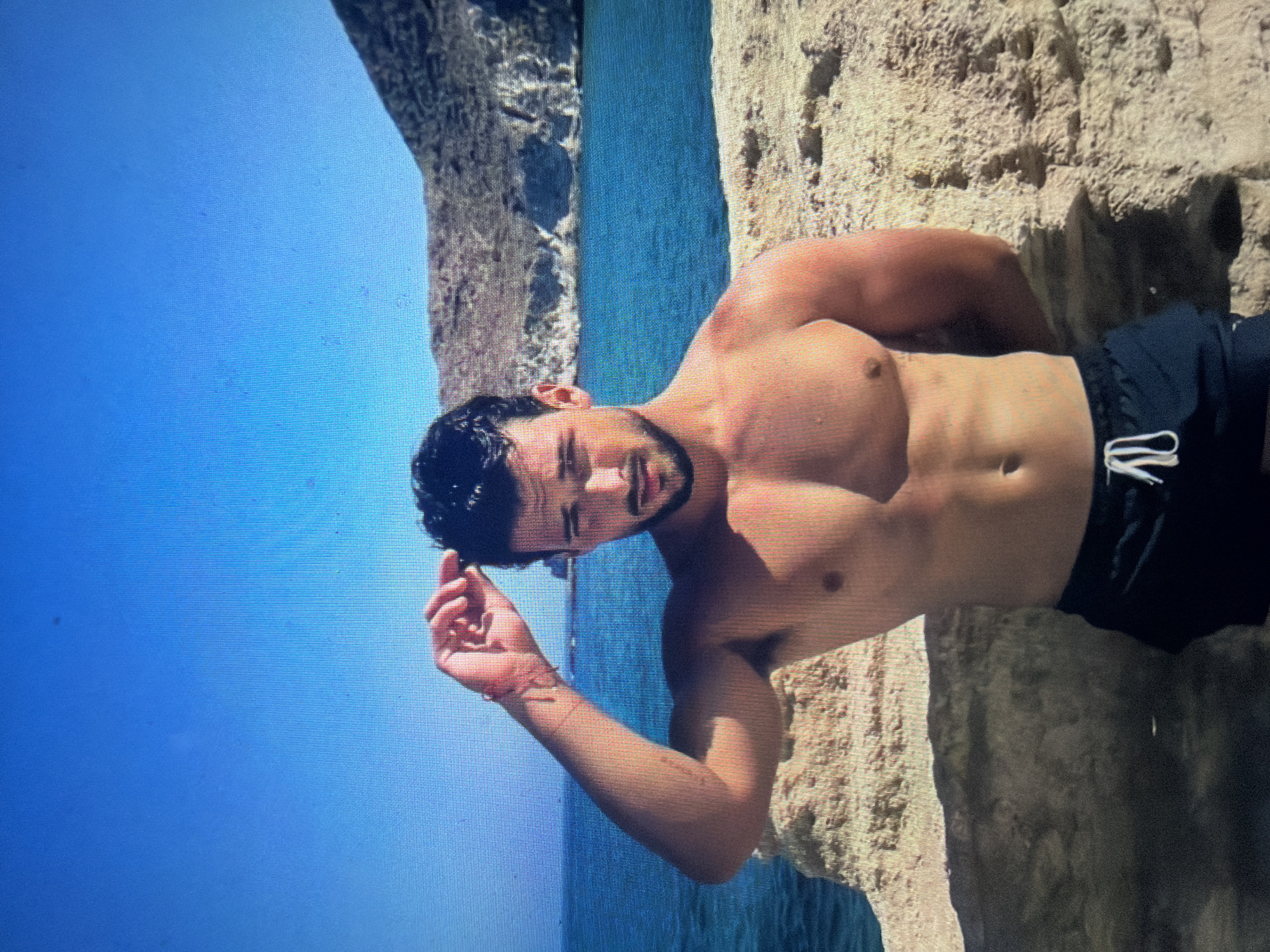 A young adult male, shirtless with a muscular build, dark hair, and a beard, stands on a sandy and rocky shore. He wears dark swim shorts with a white drawstring. His right arm is raised, with his hand touching his wet hair, while his left arm is bent behind his back. The background features a vibrant blue sea or ocean, with a distant coastline of rocky hills and cliffs under a clear blue sky. The immediate foreground consists of light-colored rock formations and sand. The scene is situated in Shumen, Bulgaria.