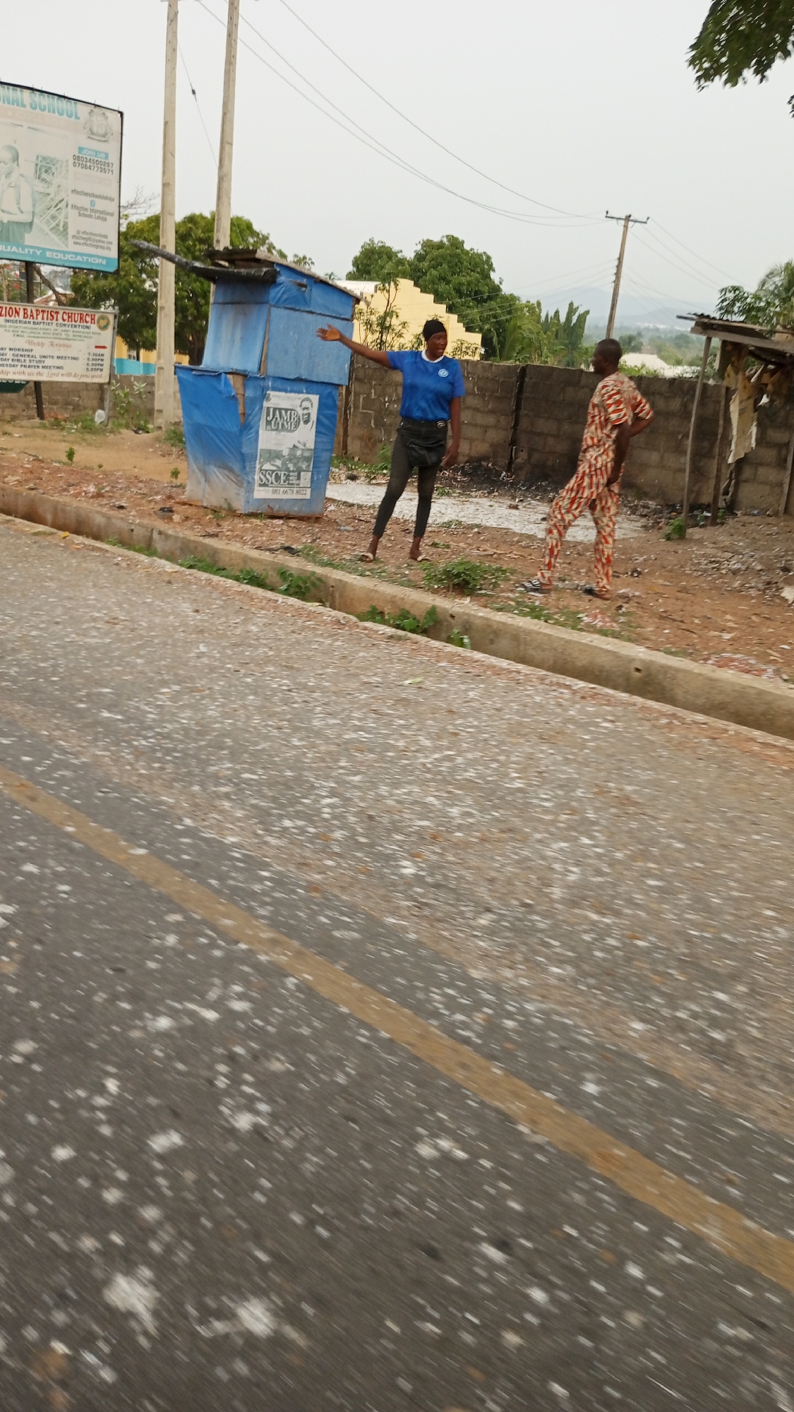 The image shows a street scene in Adankolo, Nigeria. A woman wearing a blue shirt and black pants stands near a makeshift blue structure with posters on it by the side of the road. The posters appear to be advertisements for education and examinations. She is gesturing with her right hand. A man in a patterned outfit stands nearby, with his hand on his hip. The road is asphalt with lane markings, and there are buildings and foliage in the background. Overhead power lines are visible. A church signboard with service schedules is also visible.