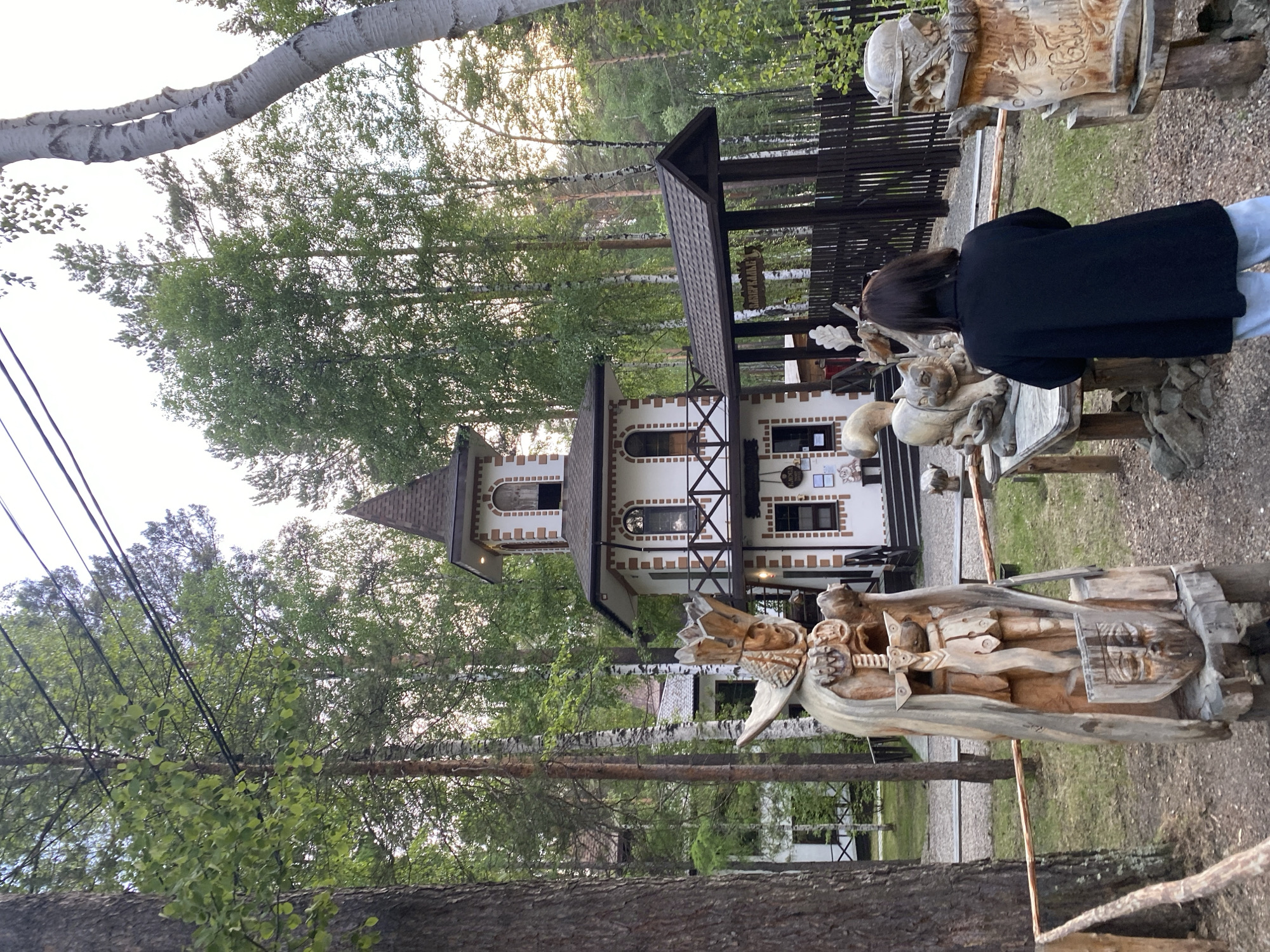 The image depicts an outdoor scene in the city of Tal'tsy, Russia, likely at an ethnographic museum or park. In the foreground, a wooden sculpture of a warrior in armor, complete with a crown, sword, and shield, stands prominently. To the right of this sculpture, a woman with dark hair, wearing a black jacket and light-colored pants, is standing, facing away from the camera and observing the scene. In the middle ground, a whimsical, multi-story building resembling a fairy-tale tower stands, with white walls, brick accents, and arched windows. A covered walkway extends from the building, leading to a dark wooden fence. Various other wooden sculptures, including a large cat-like creature and an owl-like bust, are scattered around the area. The background is dominated by tall, slender trees, characteristic of a forest or wooded park, with a bright, overcast sky overhead. The overall impression is of a rustic, fantastical, and culturally rich environment.