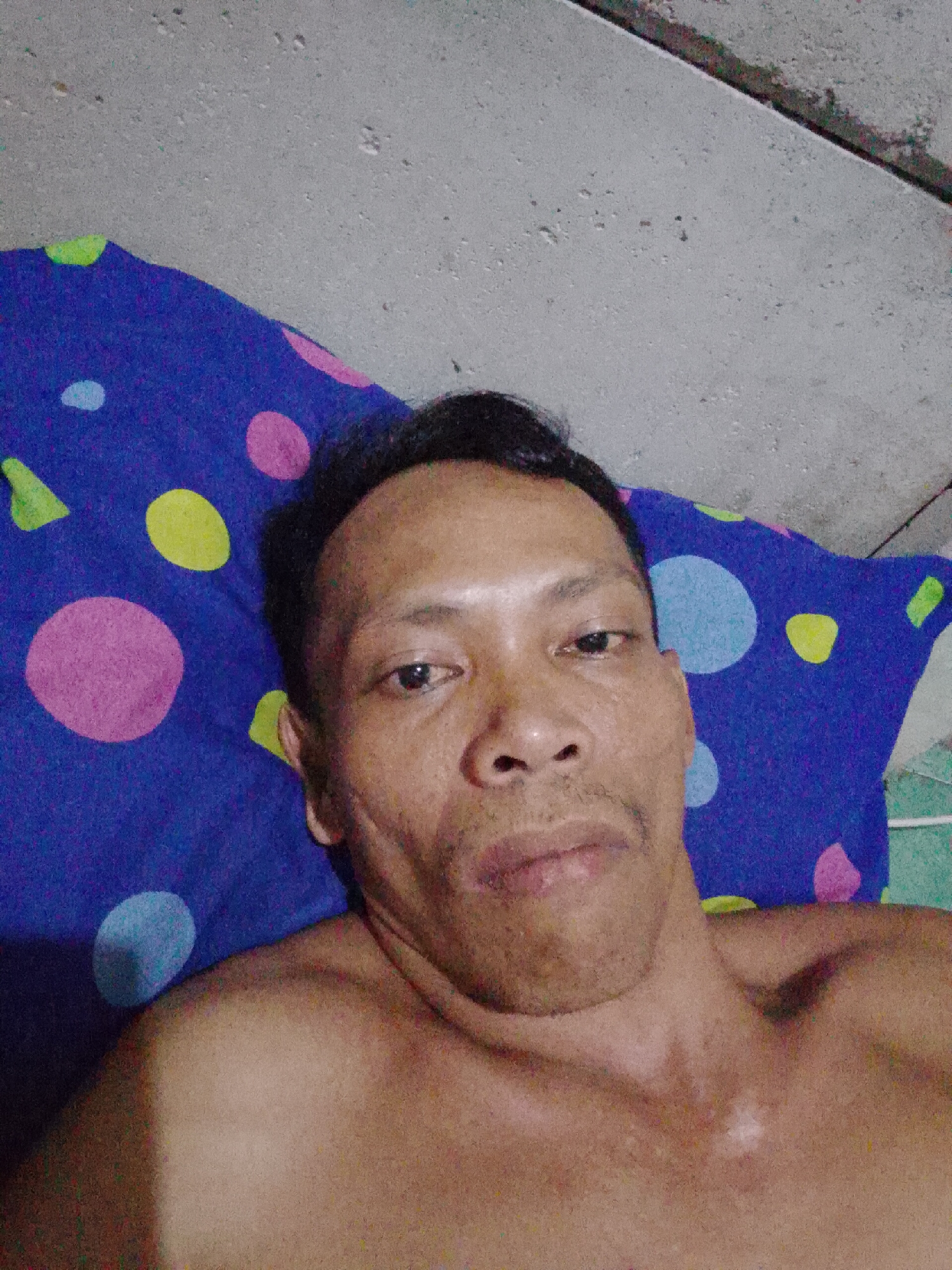 The image shows a shirtless man lying on a blue pillow with colorful polka dot patterns. He is lying on his back, looking directly at the camera. The background is a plain, light-colored wall. The photo was taken in Bukit Ketok, Indonesia.