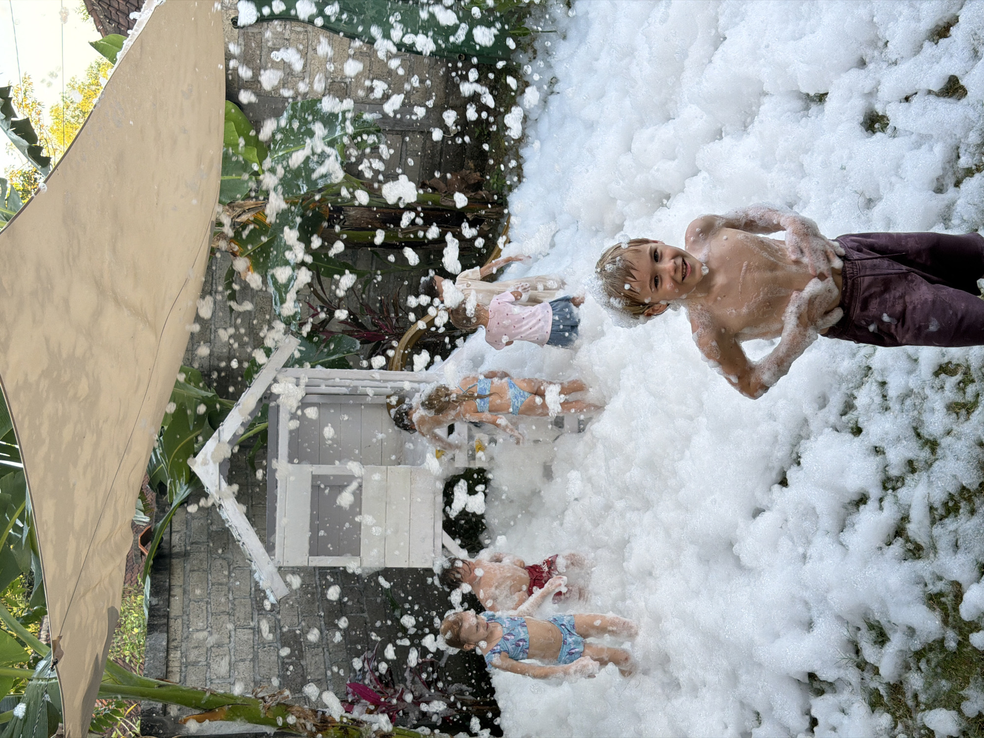 This overhead image captures a lively outdoor foam party, with several children playing enthusiastically in a vast expanse of white foam. The setting appears to be a tropical garden or backyard, indicated by the lush green foliage, including large banana-like leaves, visible around the edges and in the background.
In the foreground, a young boy, shirtless and wearing dark pants, is largely covered in foam, smiling directly at the camera with an expression of joy. Further back, several other children are visible amidst the foam. One child wears a pink top and shorts, another a light-colored garment, and two others are in blue and white patterned swimsuits. They are all submerged to varying degrees in the foam, engaged in playful activity.
To the left of the frame, a small, white playhouse or structure with visible windows and roof details is partially obscured by the foam. A large, beige triangular shade sail extends from the upper left, providing cover from the bright daylight. The ground beneath the foam, where visible, appears to be made of stone or brick paving. The overall atmosphere is one of carefree fun and excitement. The bright lighting suggests it is daytime, and the tropical vegetation aligns with the provided location context of Kuta Selatan, Indonesia. There is no visible text in the image.