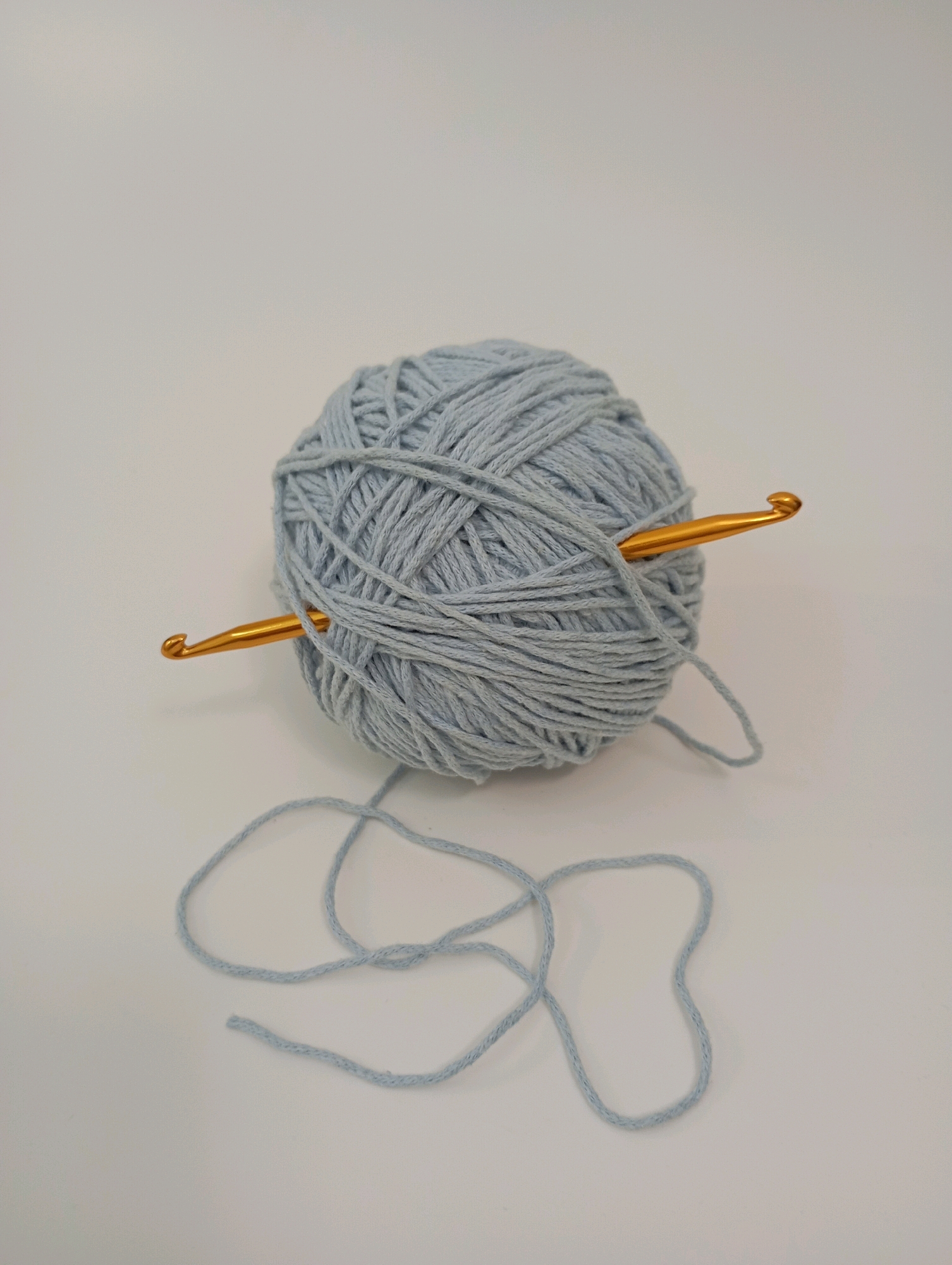 A ball of light blue yarn with a gold crochet hook inserted through it sits on a white surface. A length of yarn trails from the ball across the surface. The image is a close-up studio shot with a plain white background. The location in Krasnodar, Russia is not specified.