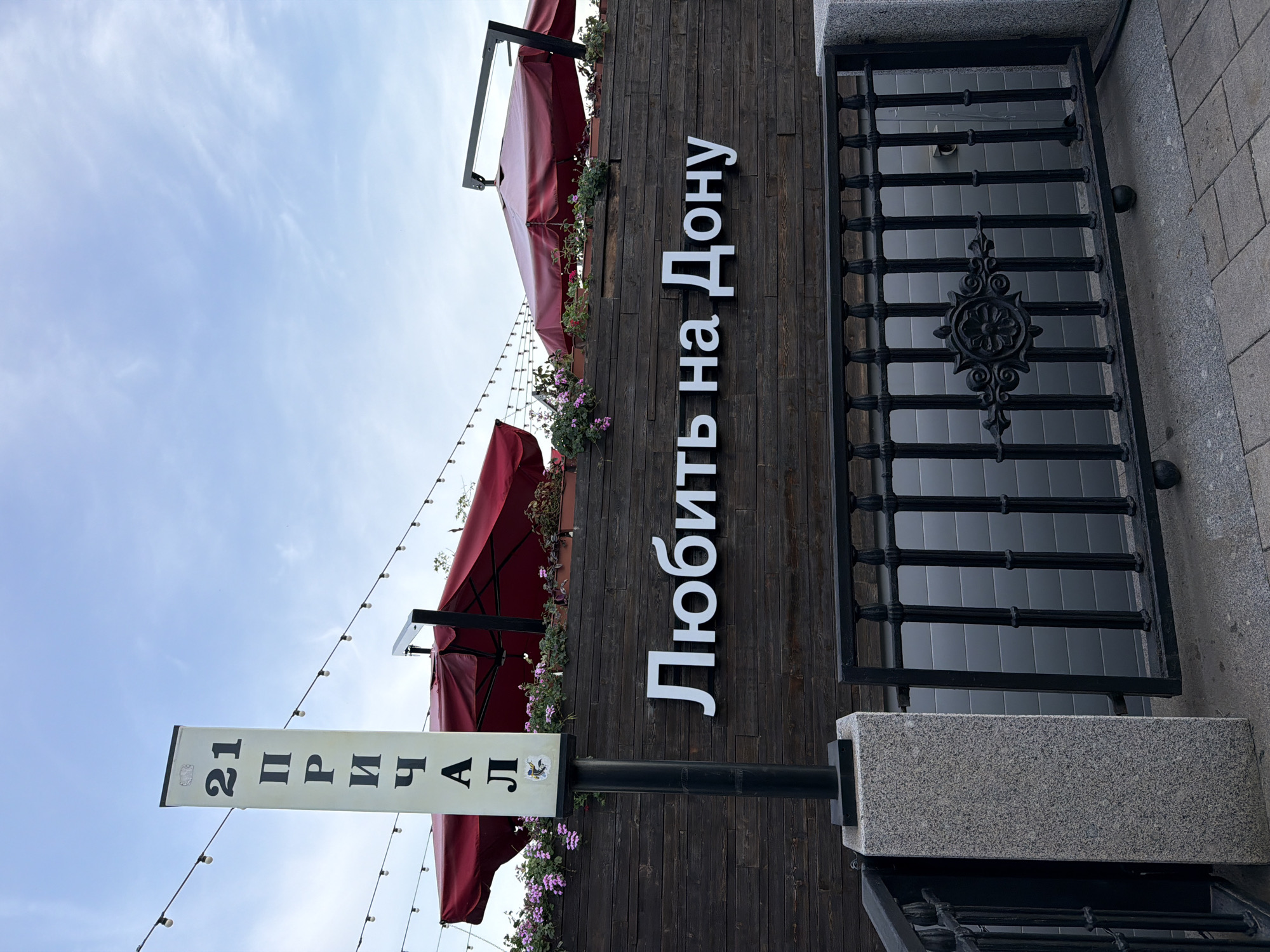 The image depicts the exterior of a business establishment in Rostov-on-Don, Russia. The sign "21 Причал" (21 Pier) is visible, indicating a restaurant or similar establishment. Above that is a wooden wall with the Cyrillic text "Любить на Дону" (Love on the Don). Red awnings and decorative floral arrangements add to the ambiance. To the right is a black metal grate or door. The sky is partly cloudy. The overall impression is that of an outdoor dining or gathering space in a city environment.