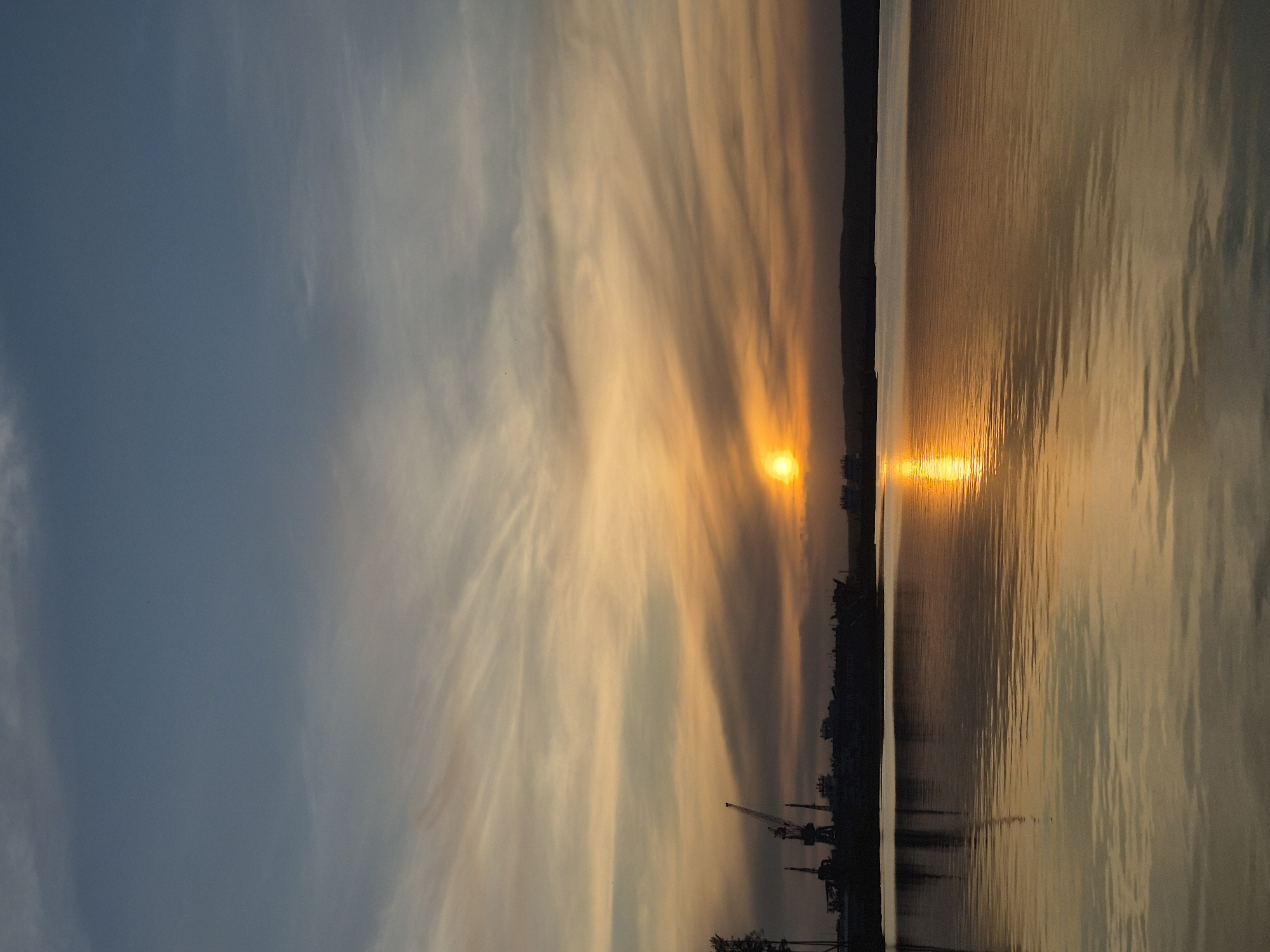The image shows a sunset over water, likely a river or lake, in Chaikovsky, Russia. The sun is near the horizon, casting a golden reflection on the water. The sky is filled with clouds, some with a wispy appearance, illuminated by the setting sun. Silhouettes of cranes and industrial structures are visible on the shoreline. The water surface has gentle ripples, reflecting the colors of the sky and sun.