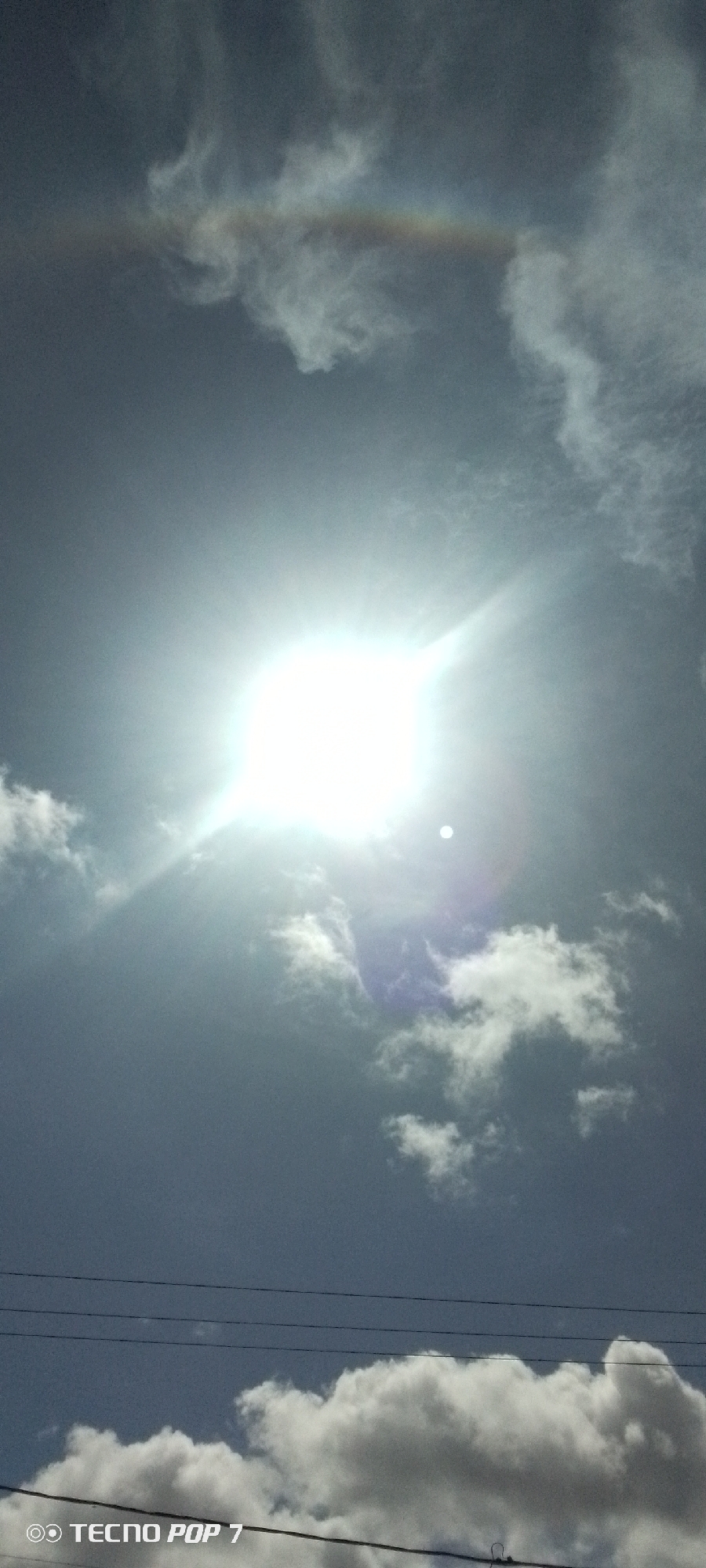 The image shows a bright sun shining through clouds, with power lines visible in the lower portion of the frame. A rainbow-like effect appears at the top of the image, possibly due to atmospheric conditions. The photograph was taken in Altagracia, Venezuela, as indicated by the EXIF data. No people or subjects are visible in the image. The main subjects are the sun, clouds, and power lines.